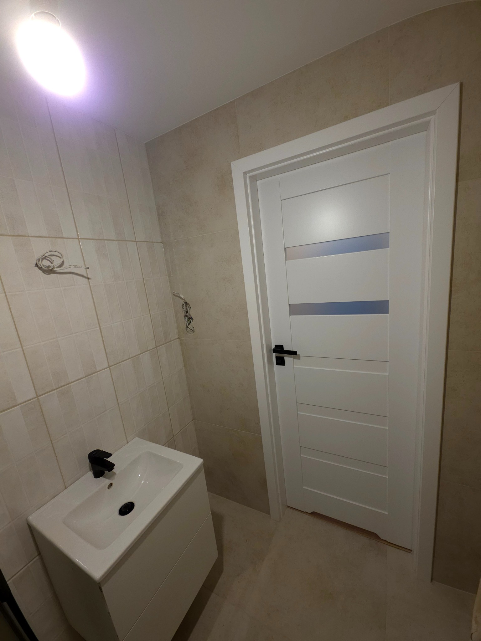 Wykończona łazienka z beżowymi płytkami, białą szafką z umywalką i czarną armaturą. Widoczne białe drzwi z przeszkleniami i wystające przewody.