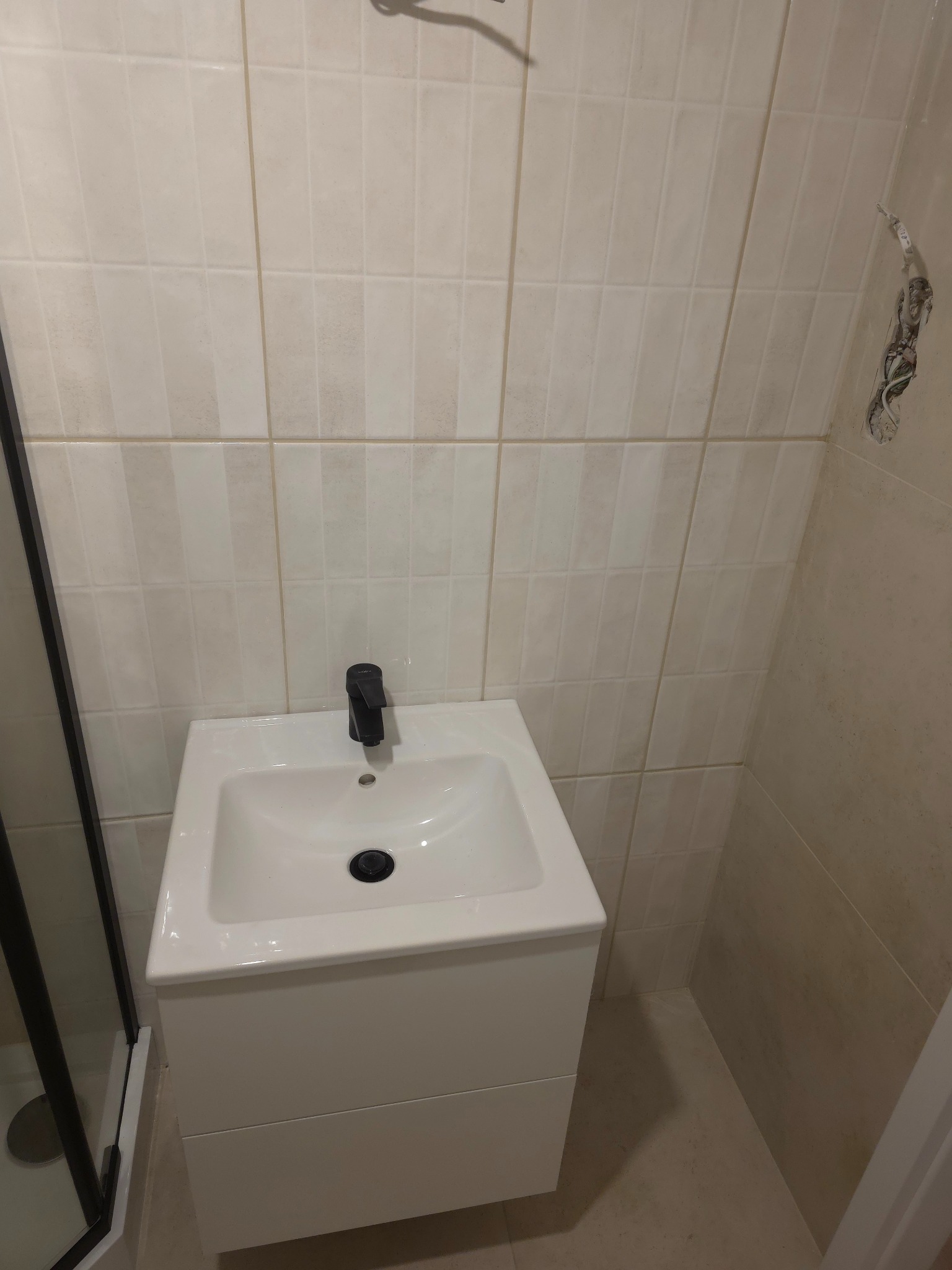 Biała umywalka z czarną baterią w wykończonej łazience z beżowymi, pionowymi płytkami. Widoczne fragmenty kabiny prysznicowej i niedokończone gniazdko elektryczne.