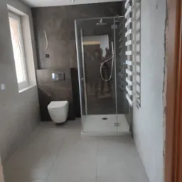 Nowoczesna łazienka po remoncie z kabiną prysznicową ze szklanymi drzwiami, wiszącą toaletą na tle ciemnobrązowej ściany i białym grzejnikiem drabinkowym, oświetlona reflektorami szynowymi.