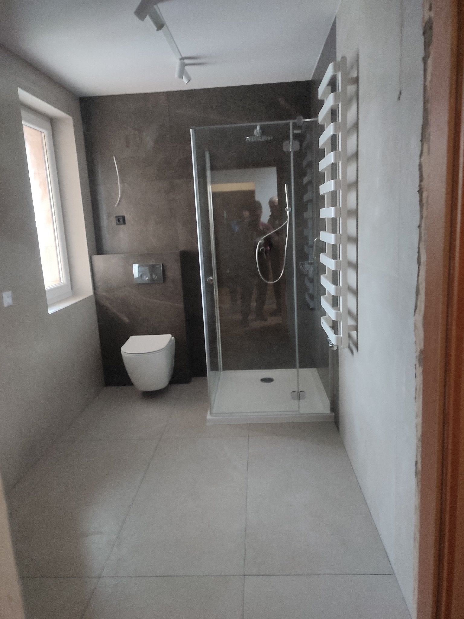 Nowoczesna łazienka po remoncie z kabiną prysznicową ze szklanymi drzwiami, wiszącą toaletą na tle ciemnobrązowej ściany i białym grzejnikiem drabinkowym, oświetlona reflektorami szynowymi.