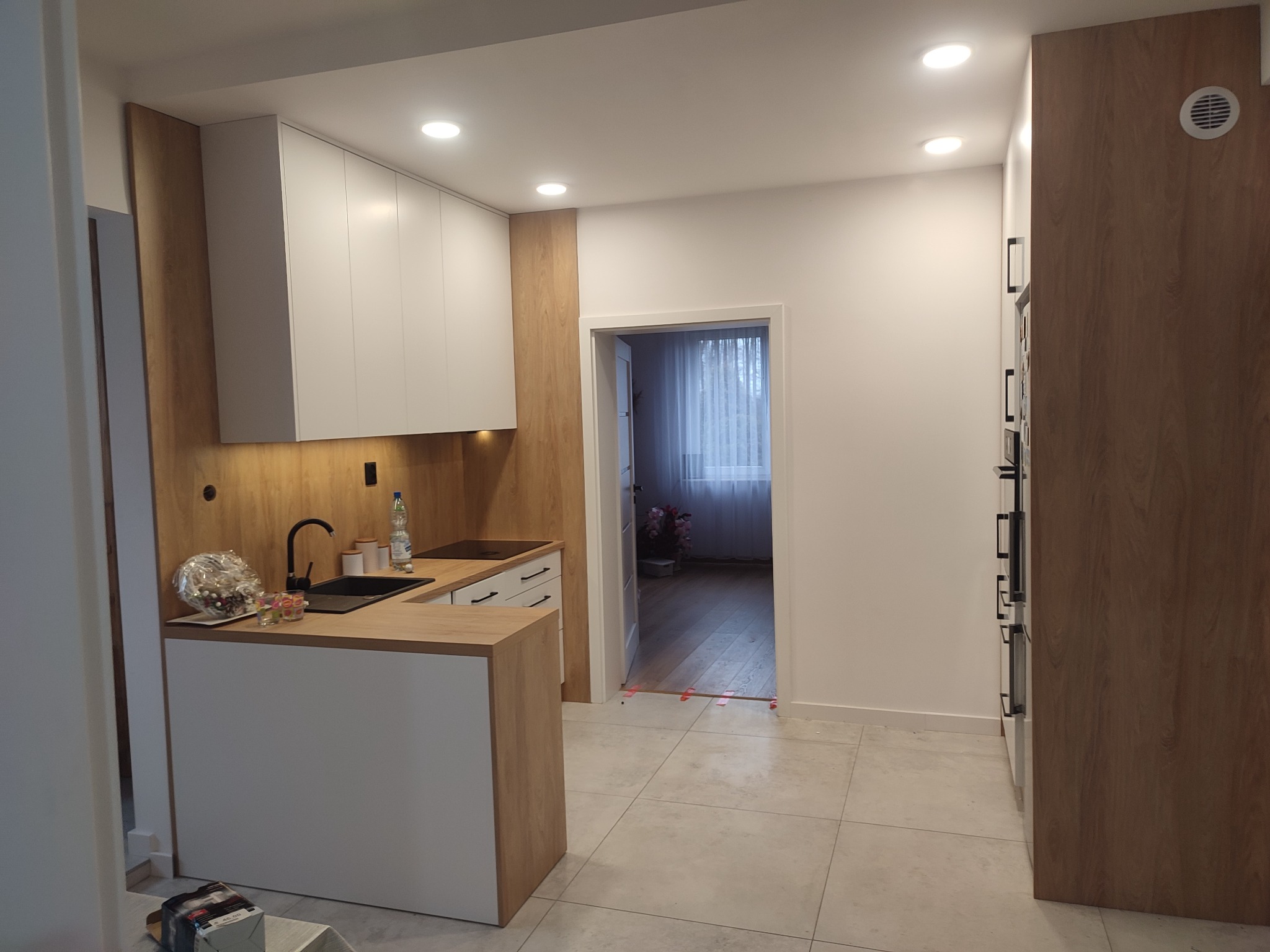 Nowoczesna kuchnia z białymi szafkami, drewnianymi akcentami i czarną armaturą, widok na sąsiedni pokój przez otwarte drzwi, na blacie ozdobny bukiet.