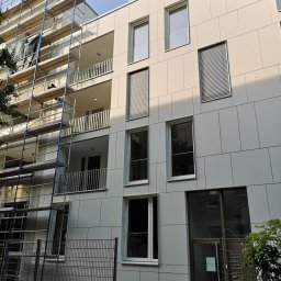 M.S.T Expo - Wielopiętrowy budynek z jasną elewacją z płyt, z widocznym rusztowaniem przy jednej ze ścian i ogrodzeniem na pierwszym planie.