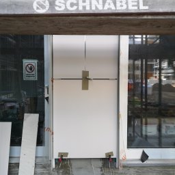 M.S.T Expo - Montaż białej płyty warstwowej w nowym budynku, widoczne narzędzia, znak zakazu wstępu na szybie, element drewniany z napisem 'SCHNABEL' u góry.