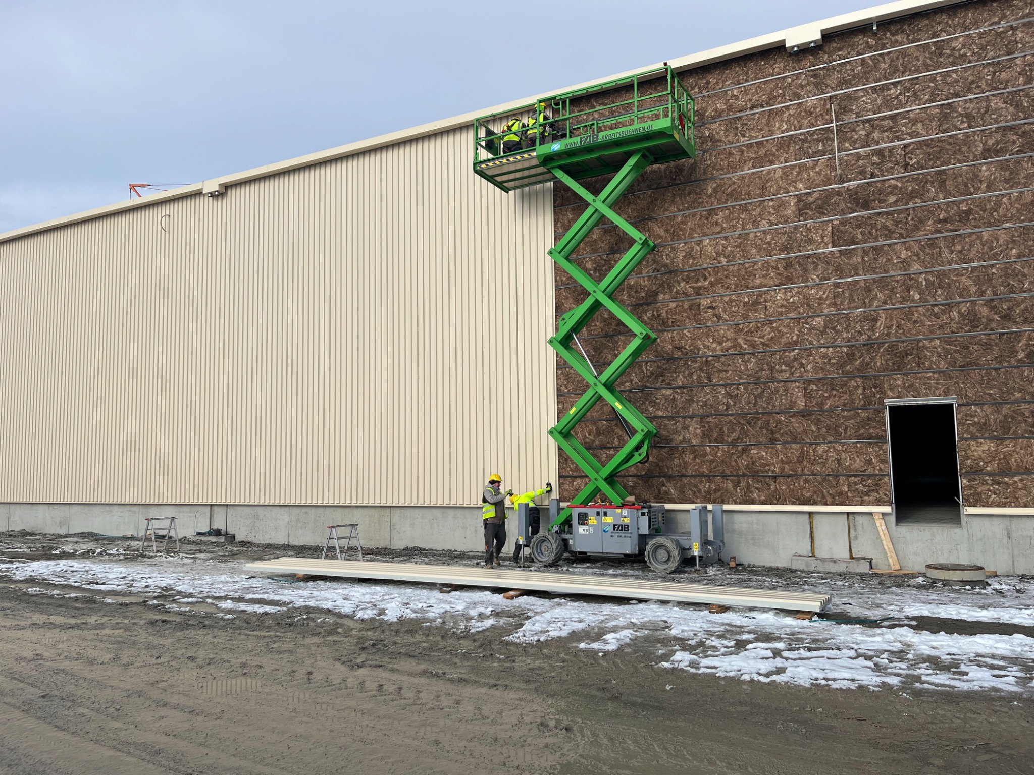 Pracownicy na podnośniku koszowym montują brązowe płyty izolacyjne na ścianie hali. Część ściany pokryta beżową blachą trapezową. Na ziemi leżą płyty warstwowe.