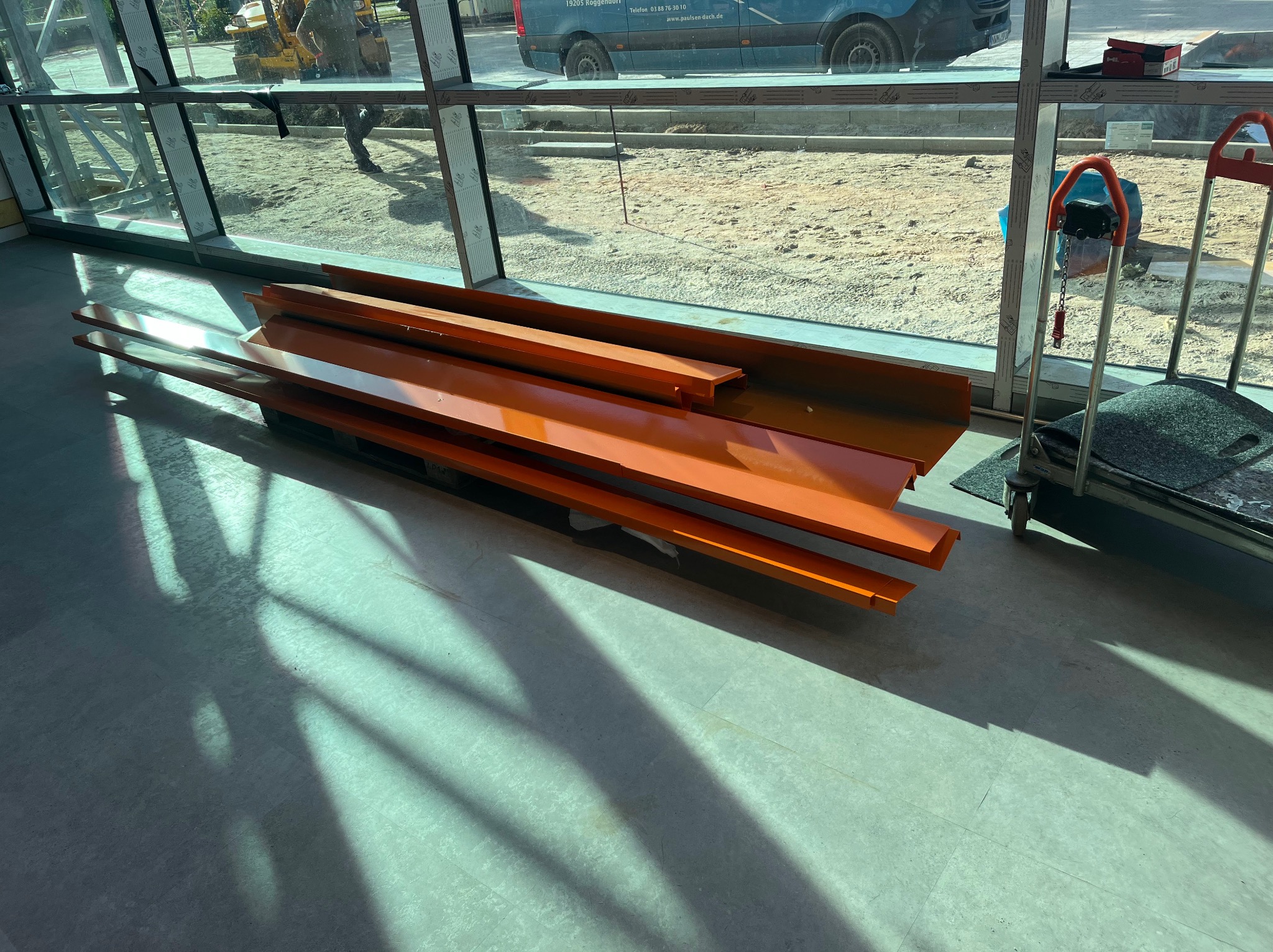 Pomarańczowe profile stalowe leżące na palecie wewnątrz budynku w trakcie budowy, widoczne przez okno maszyny budowlane i samochód dostawczy.