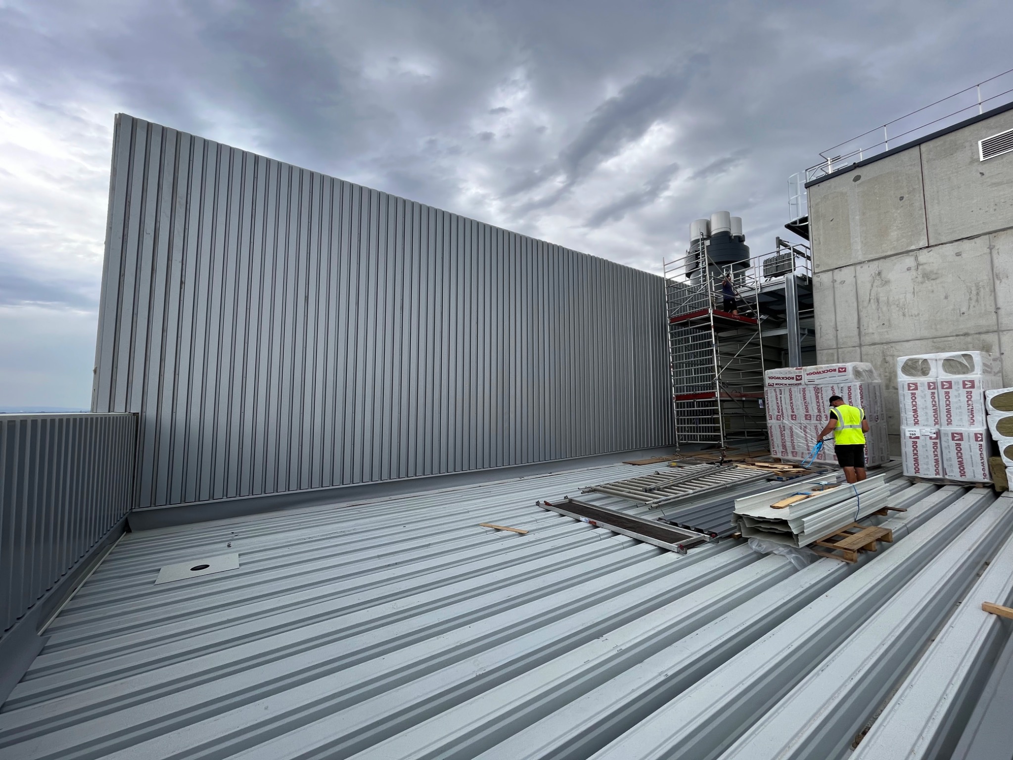 Widok z góry na plac budowy hali z częściowo zamontowanymi panelami ściennymi z blachy trapezowej, paletami materiałów izolacyjnych Rockwool i pracownikiem w żółtej kamizelce na rusztowaniu.