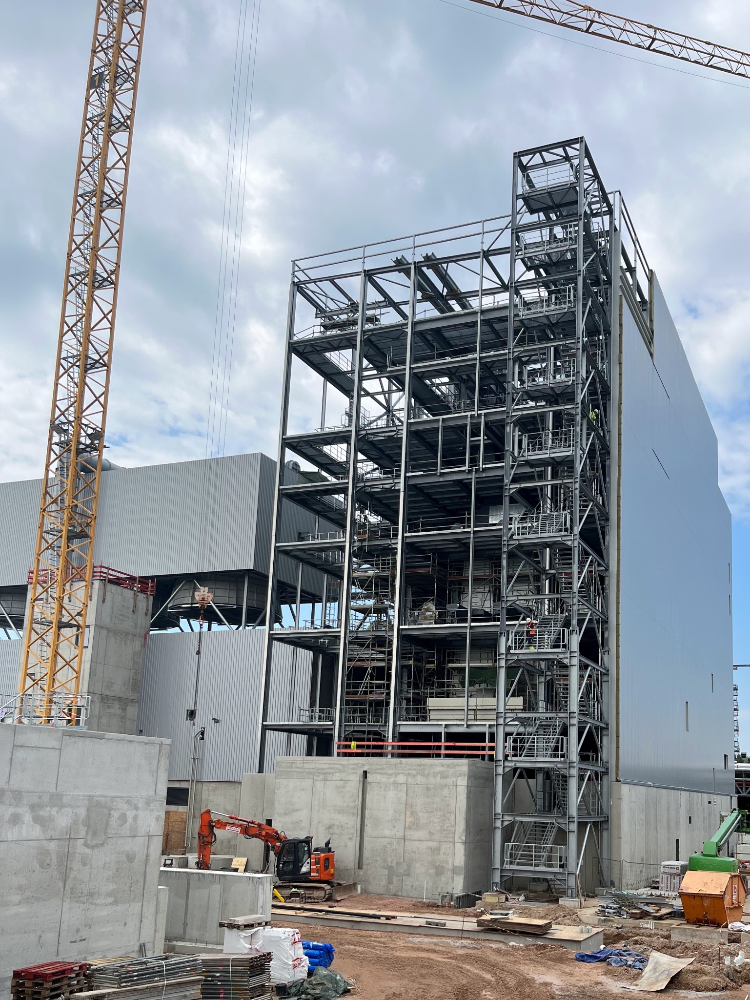 Wznoszenie stalowej konstrukcji budynku przemysłowego z widocznym szkieletem, częściowo obłożonymi płytami. Na pierwszym planie pomarańczowa koparka i elementy betonowe. W tle wysoki żuraw budowlany.