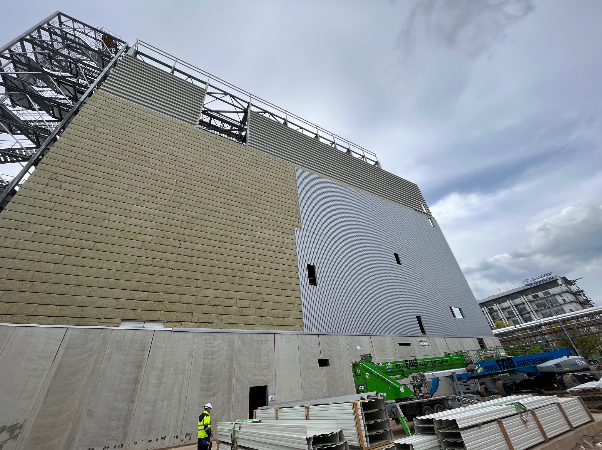 Wznoszenie ściany hali przemysłowej z widoczną warstwą izolacji i metalową elewacją, pracownik w odblaskowej kamizelce na tle palet z materiałami budowlanymi i podnośnika koszowego.