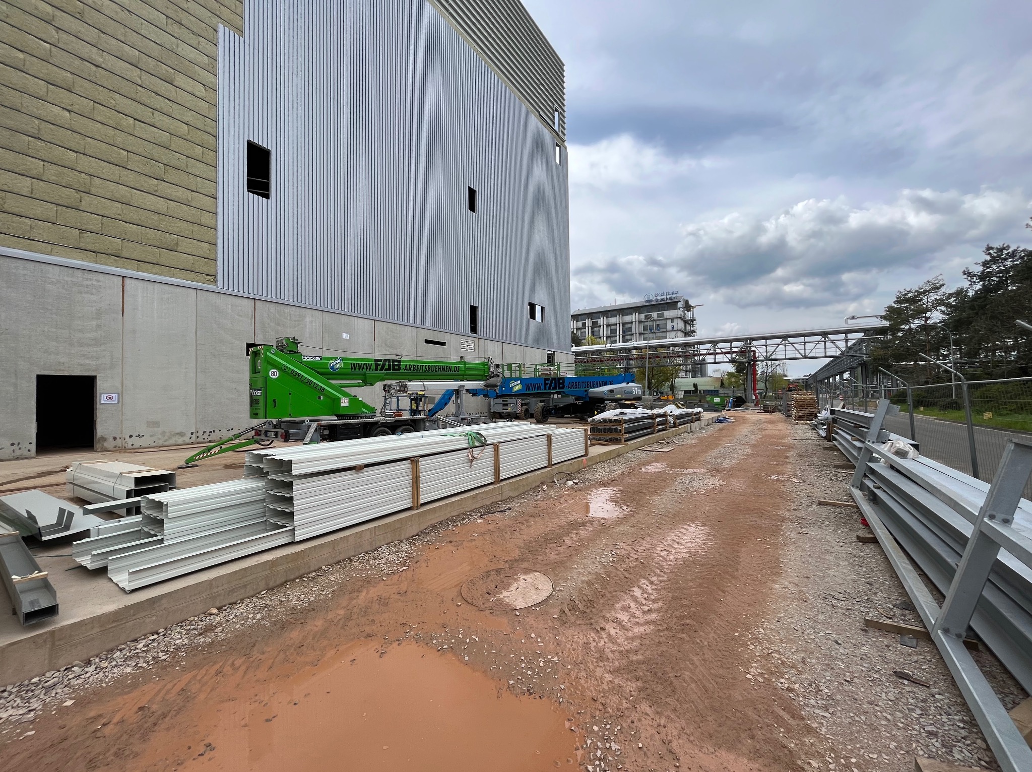 Budowa hali: widok na fragment ściany z płyt warstwowych, przygotowane elementy konstrukcyjne, podnośniki koszowe i błotniste podłoże.