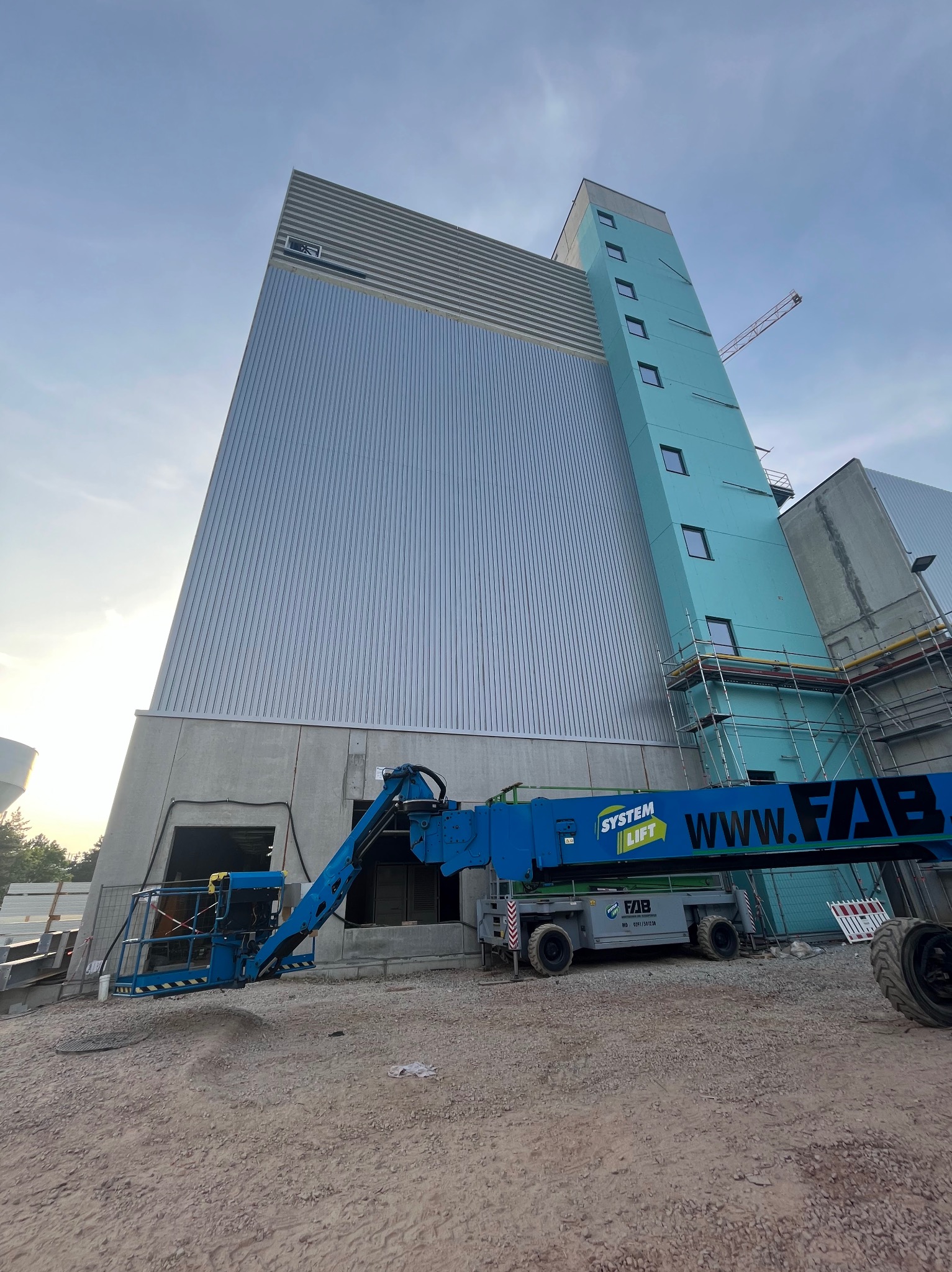 Wysoka hala przemysłowa z płyt warstwowych w trakcie budowy, widoczny podnośnik koszowy System Lift i fragment rusztowania przy jasnoniebieskiej wieży z oknami.