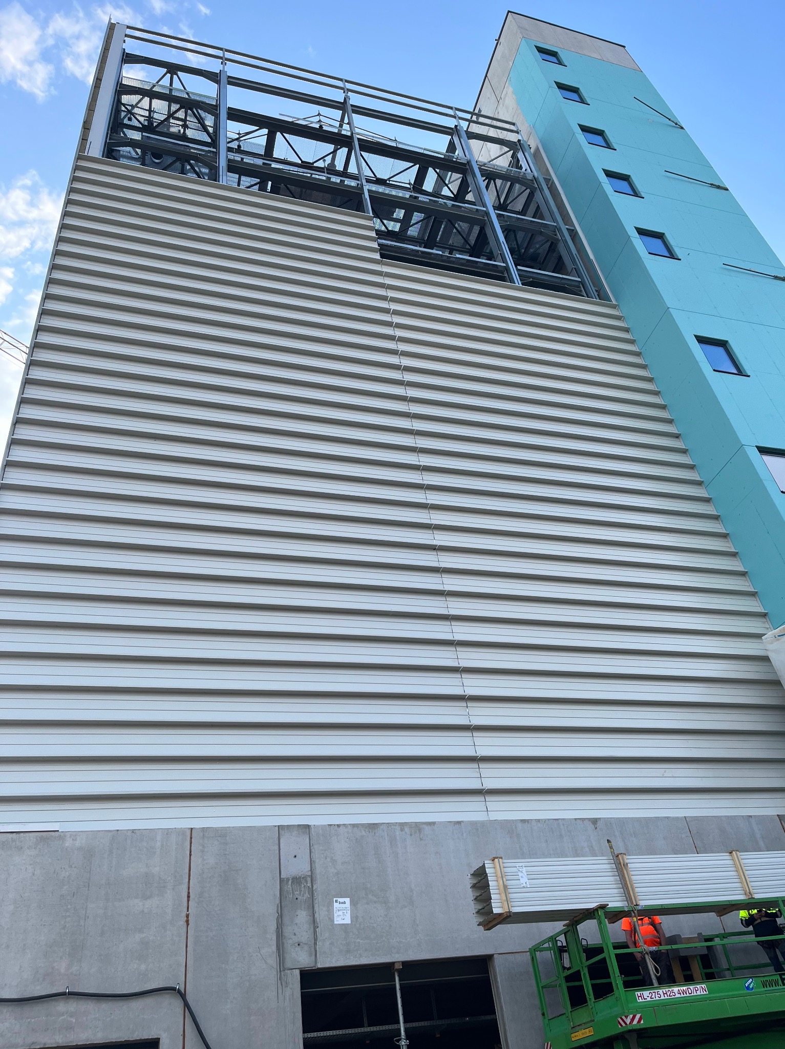 Wznoszący się budynek w trakcie montażu elewacji z płyt warstwowych, widoczna stalowa konstrukcja szkieletowa i podnośnik z paletą płyt na tle błękitnego nieba.