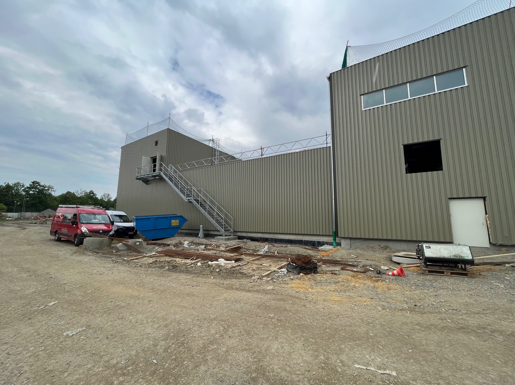 Budynek przemysłowy w trakcie budowy z widoczną konstrukcją stalową, schodami ewakuacyjnymi i przygotowanym podłożem. Na pierwszym planie gruz, zbrojenia i zaparkowane busy.