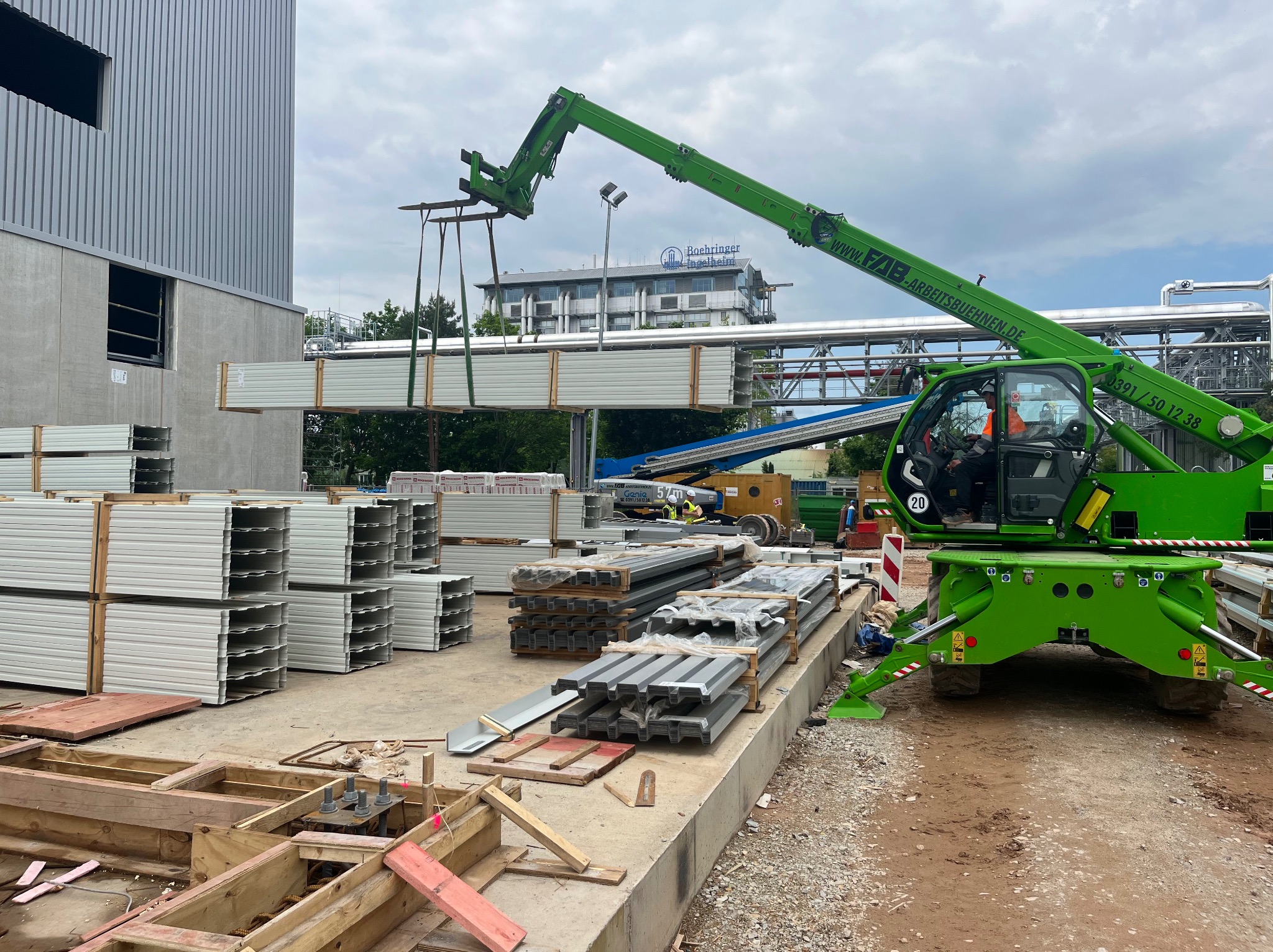Zielony podnośnik teleskopowy transportuje pakiet białych paneli na placu budowy hali stalowej. Widoczne fundamenty i przygotowane pakiety paneli.