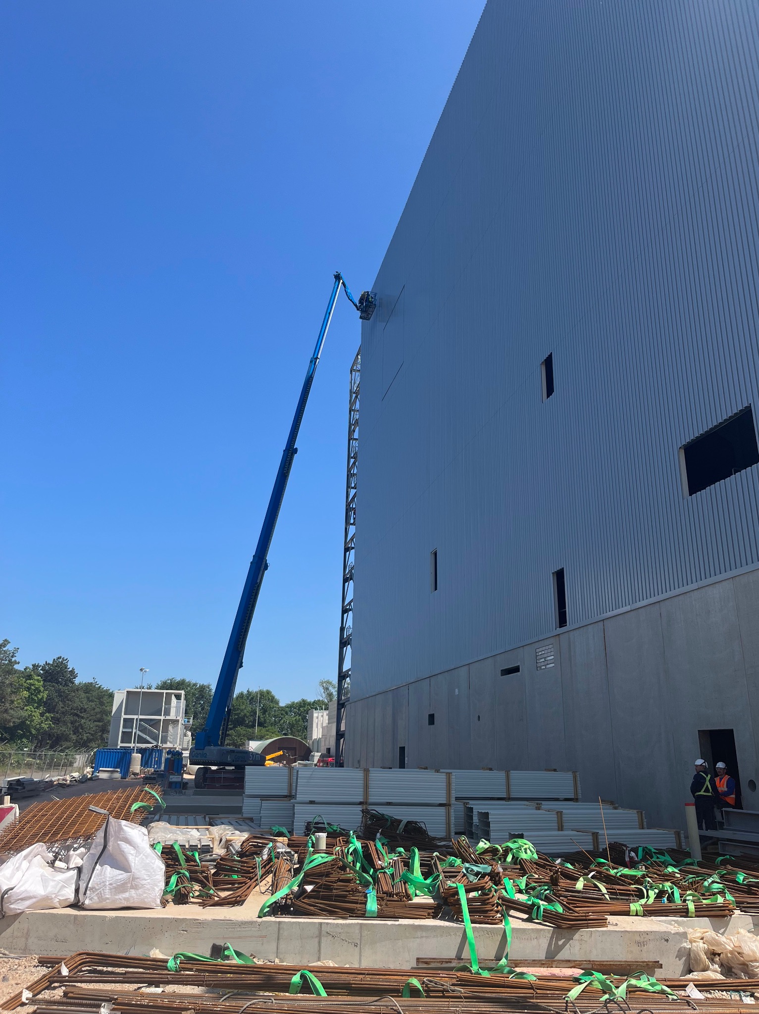 Montaż elewacji hali przemysłowej z użyciem podnośnika koszowego, widoczne przygotowane elementy konstrukcyjne i dwóch pracowników w odblaskowych kamizelkach.