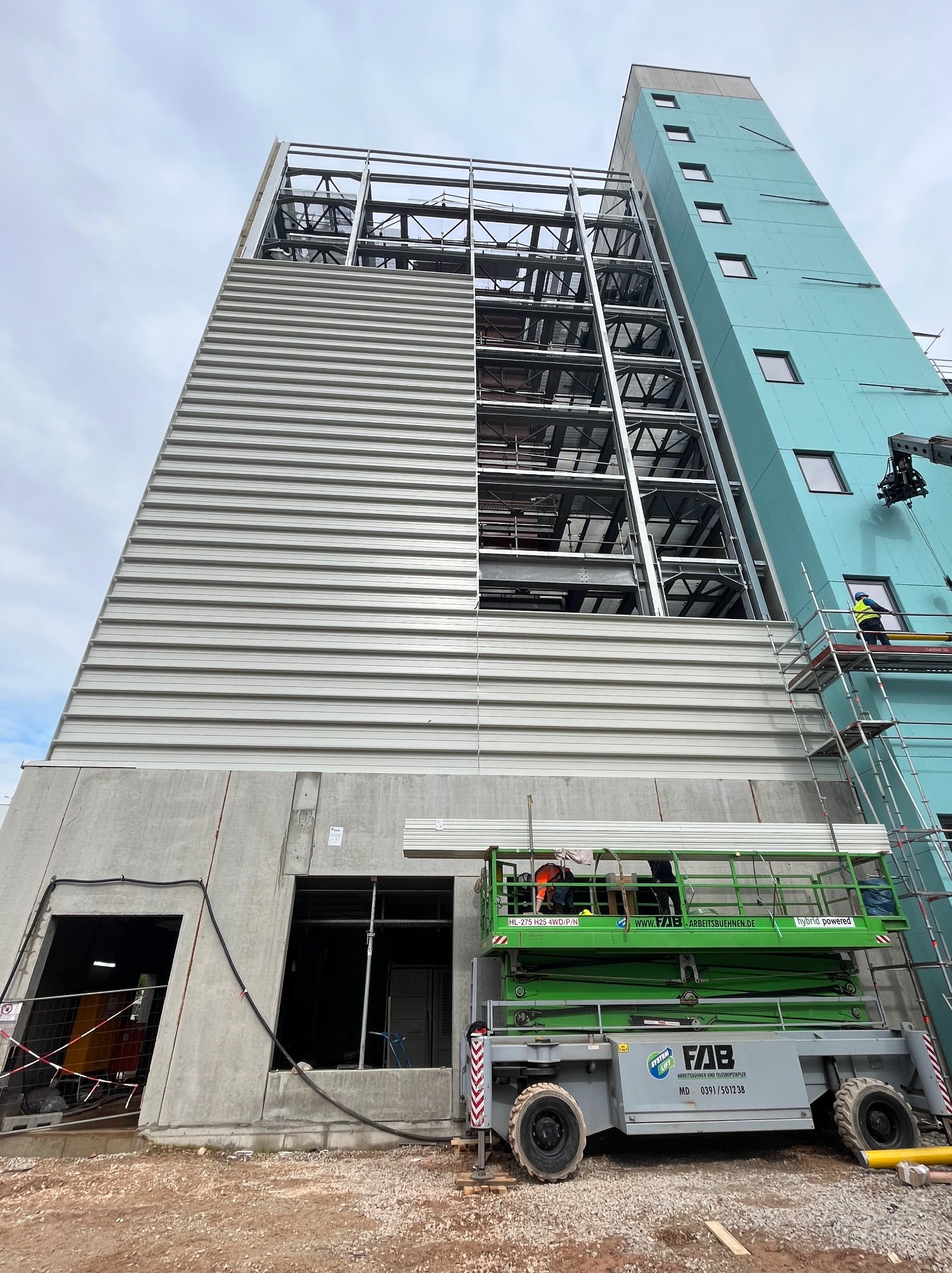 Wysoki budynek w trakcie budowy, widoczna stalowa konstrukcja i częściowo zamontowane szare płyty warstwowe, pracownicy na podnośniku koszowym i rusztowaniu, błękitna ściana budynku w tle.