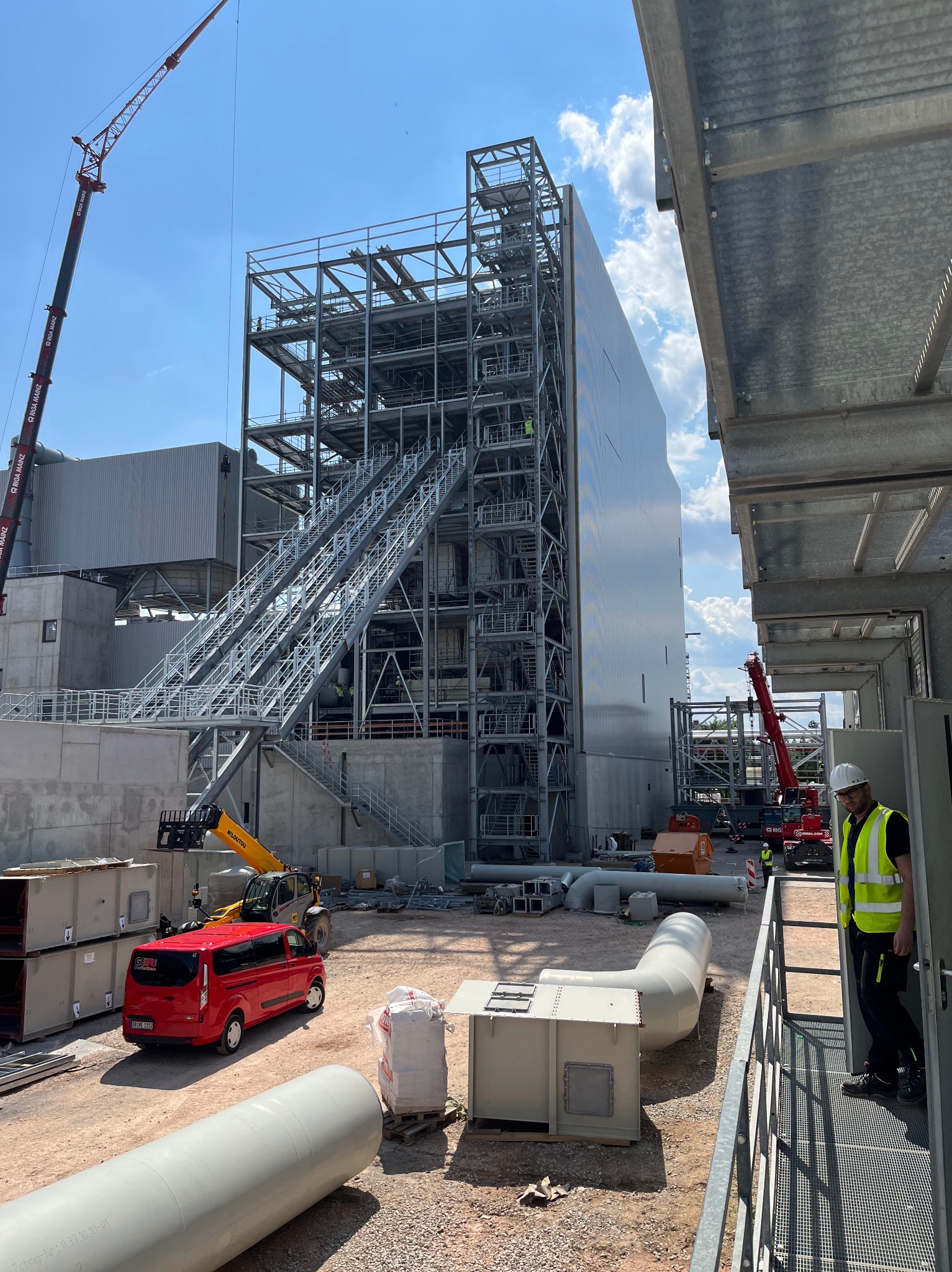 Budowa stalowej hali z widoczną konstrukcją szkieletową, schodami technicznymi, dźwigiem, czerwoną furgonetką, elementami wentylacji i pracownikiem w kamizelce odblaskowej na pierwszym planie.