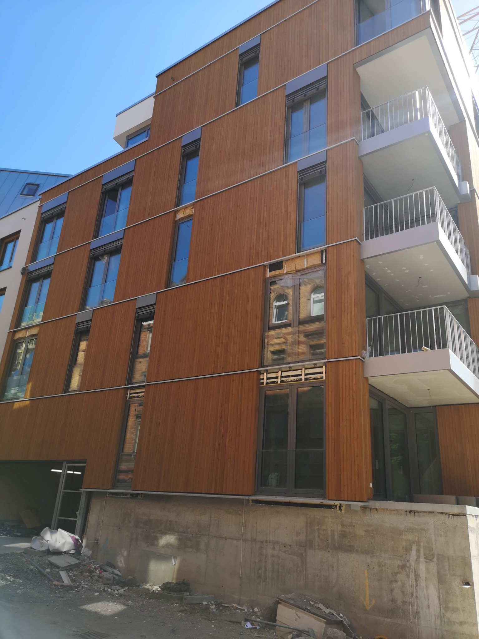 Wielopiętrowy budynek z drewnianą elewacją w trakcie budowy, widoczne niezabezpieczone otwory okienne i materiały budowlane na placu budowy.
