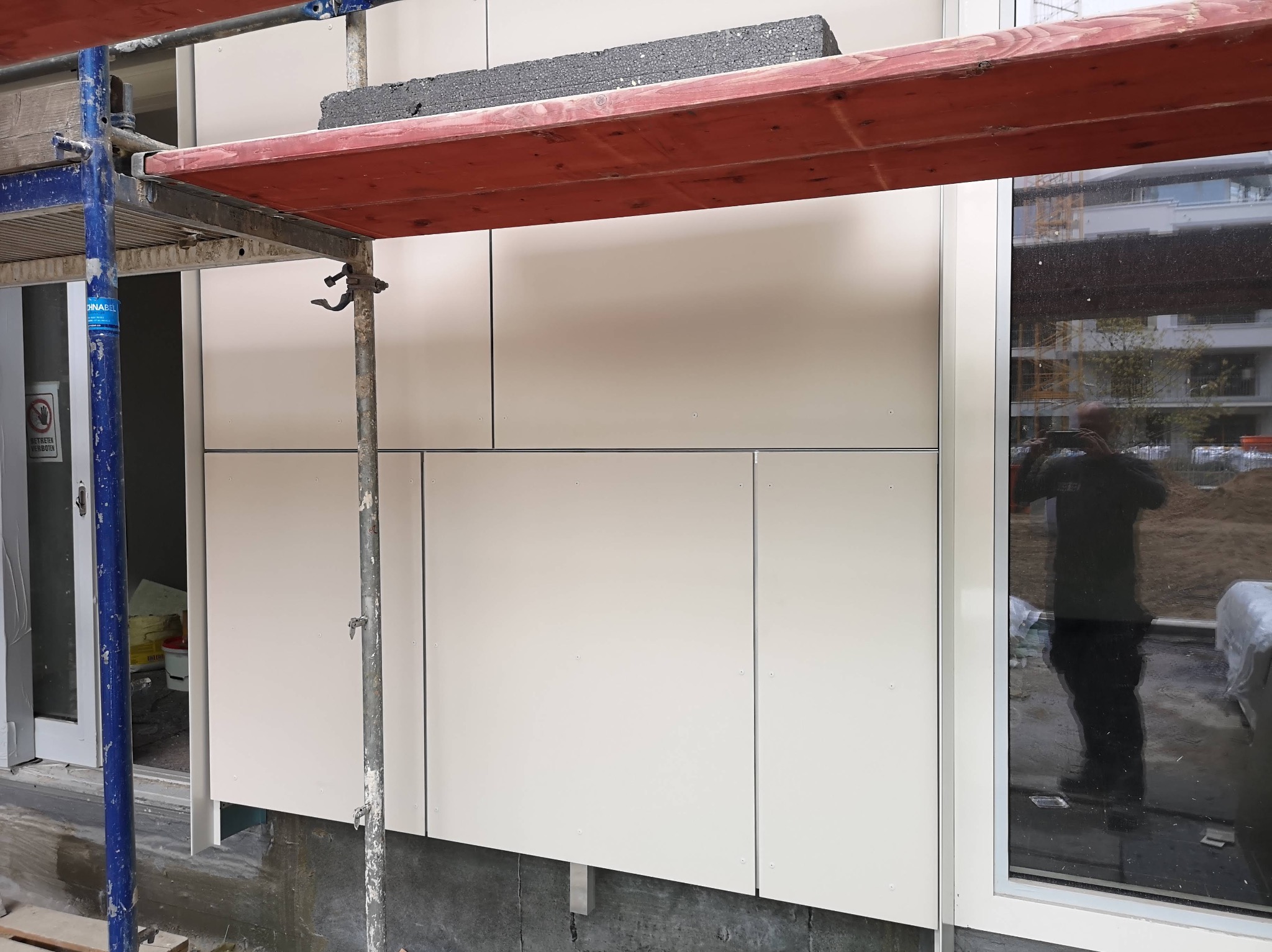 Fragment elewacji budynku z jasnych płyt, widoczne rusztowanie i odbicie osoby w oknie.