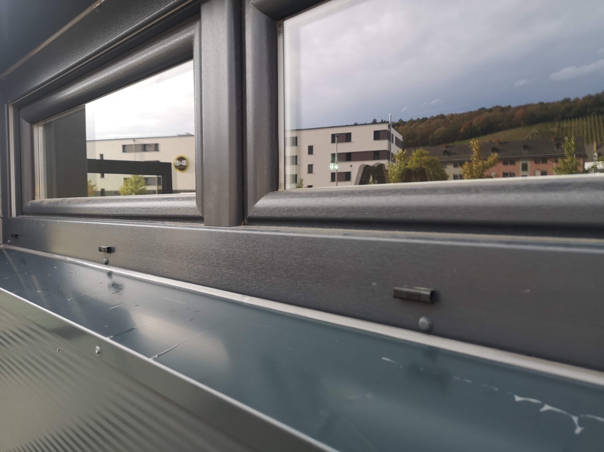 Szare okno z widokiem na budynek i zalesione wzgórze w tle, uchwycone z bliska z niskiego kąta.