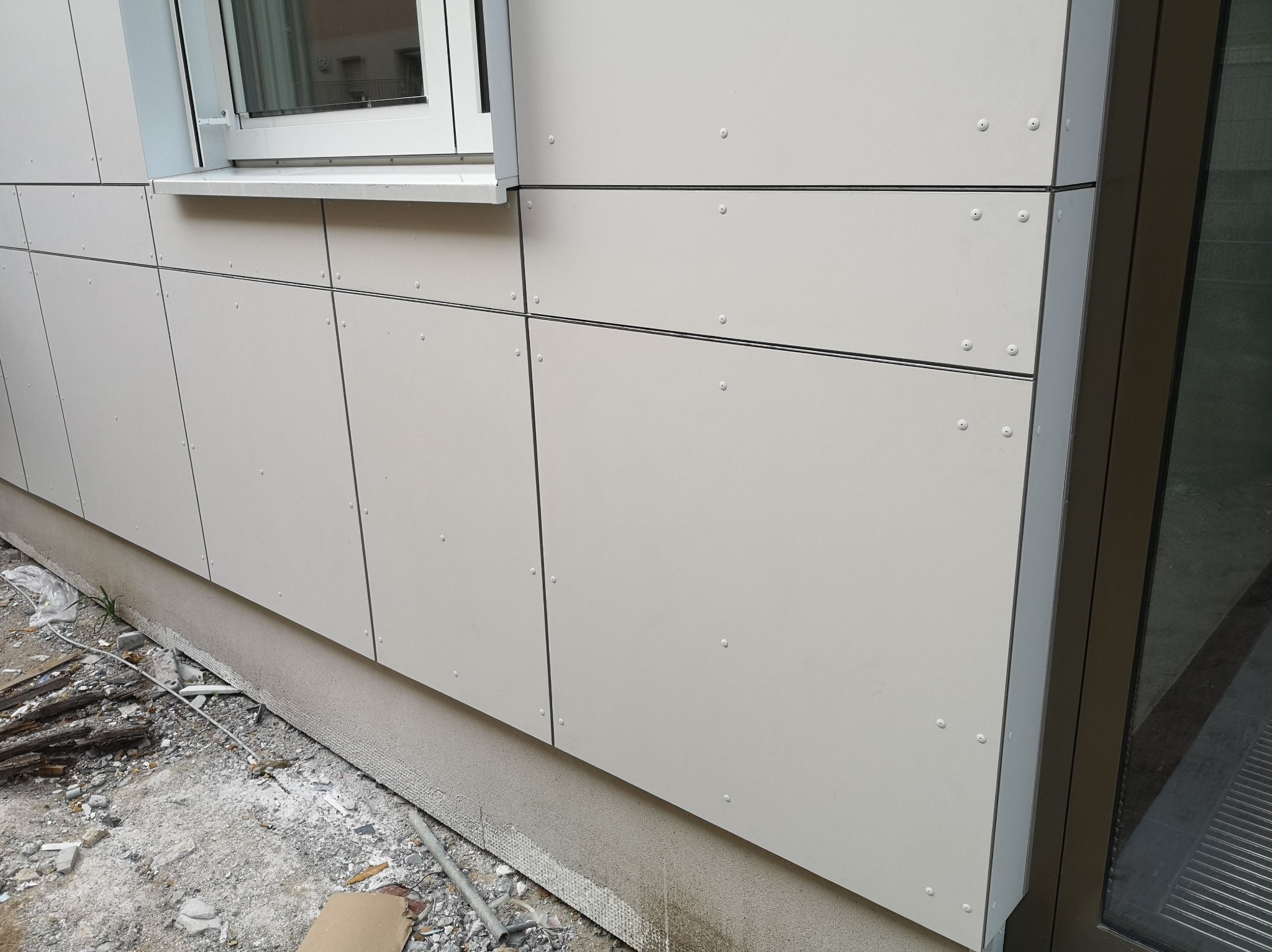 Fragment elewacji budynku z jasnych płyt warstwowych mocowanych na nity, widoczny fragment okna i drzwi, teren budowy u dołu.