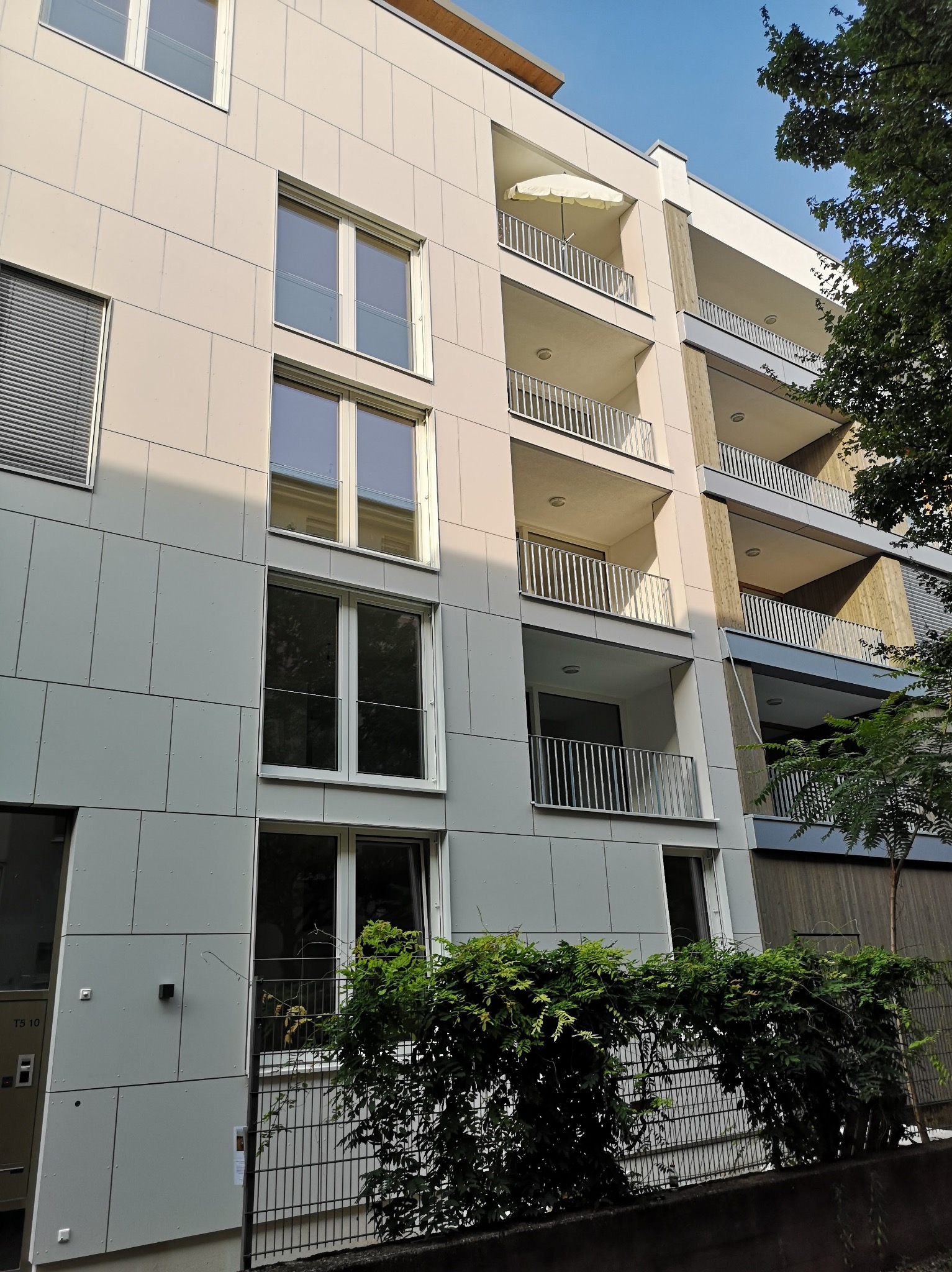 Wielopiętrowy budynek mieszkalny z balkonami, wykończony jasnymi panelami elewacyjnymi, widok z dołu, zieleń roślin w tle.