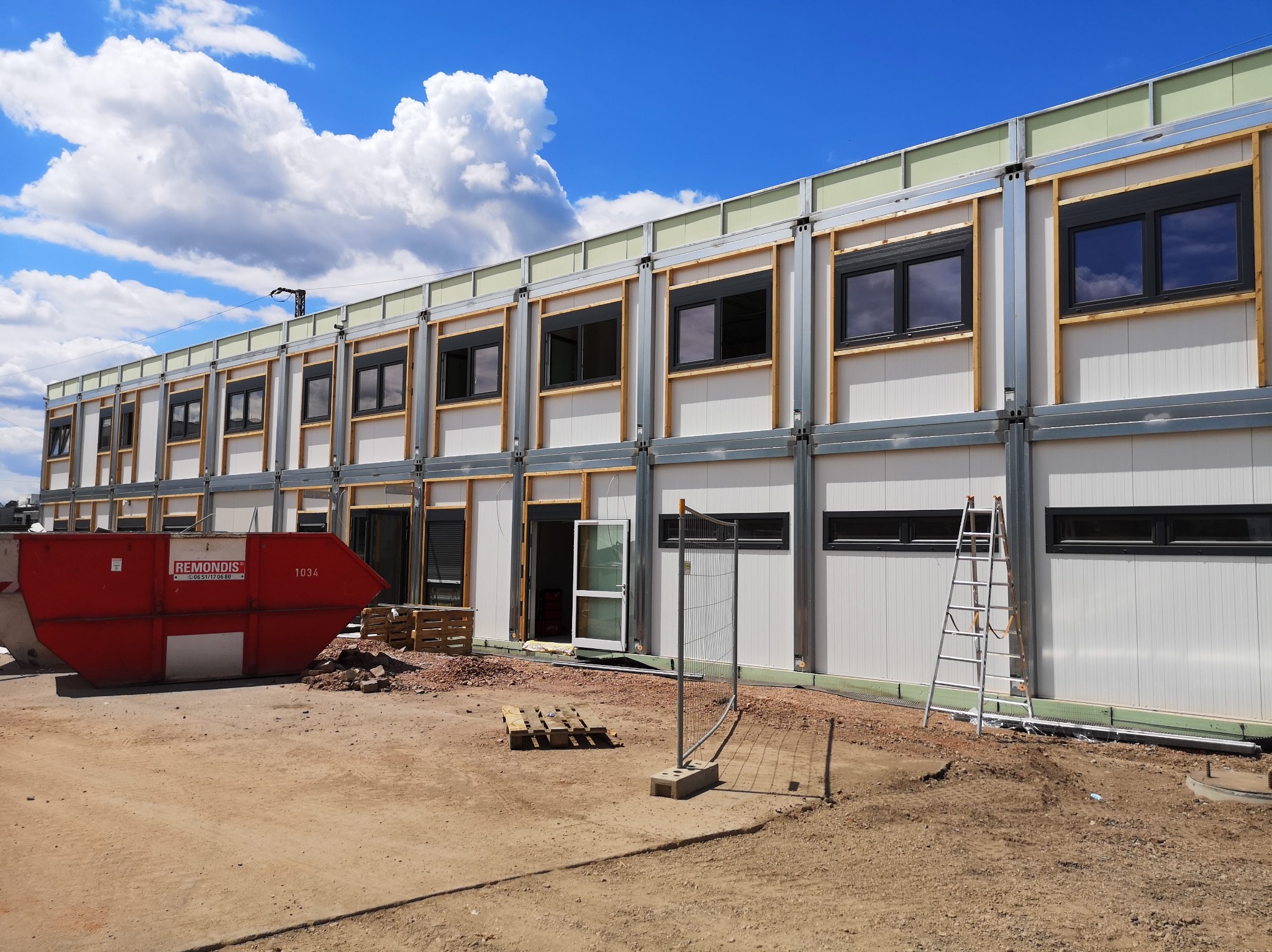 Dwukondygnacyjny budynek modułowy w trakcie montażu, widoczne stalowe ramy, białe płyty warstwowe, okna z czarnymi ramami, czerwony kontener na gruz i drabina oparta o ścianę
