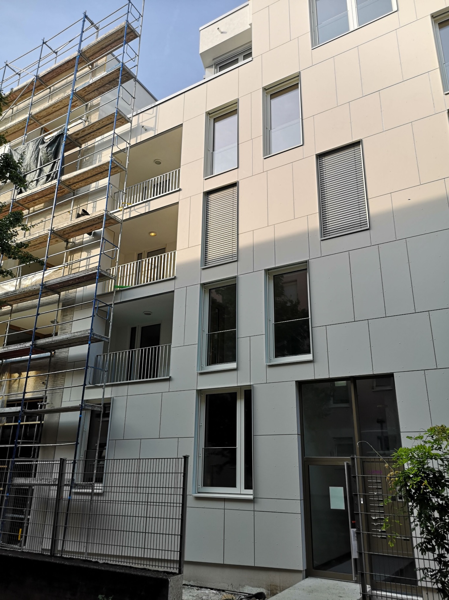 Wielopiętrowy budynek z jasną elewacją z płyt, z widocznym rusztowaniem przy jednej ze ścian i ogrodzeniem na pierwszym planie.