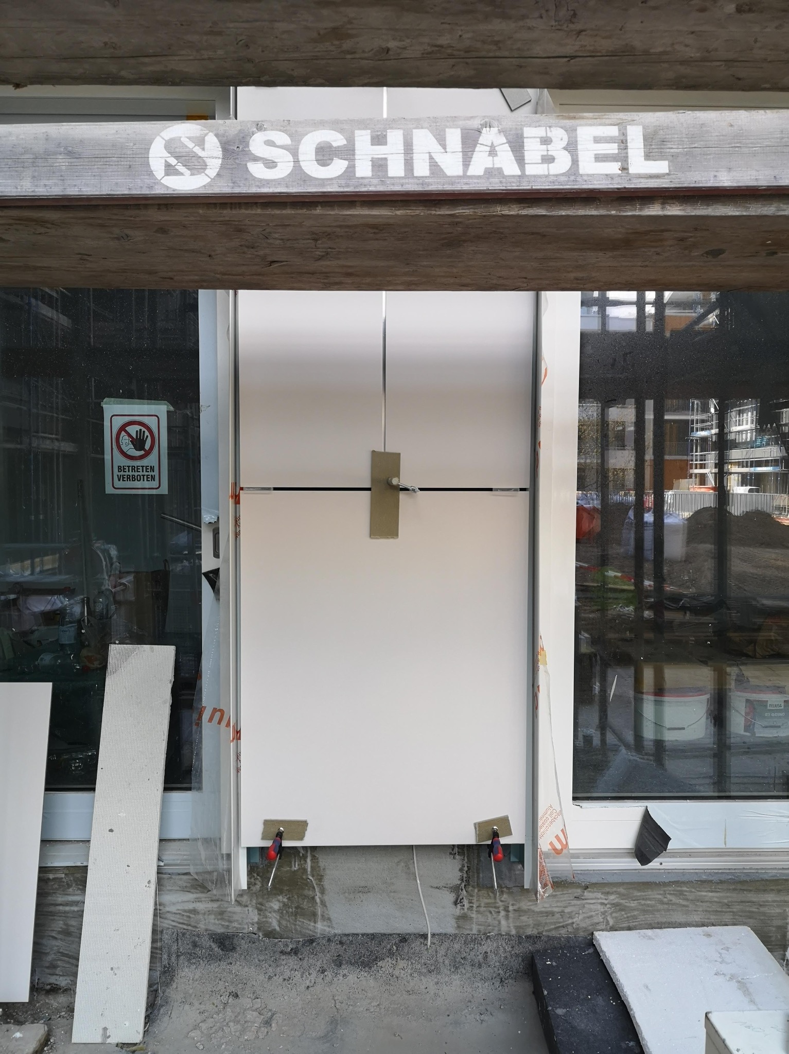 Montaż białej płyty warstwowej w nowym budynku, widoczne narzędzia, znak zakazu wstępu na szybie, element drewniany z napisem 'SCHNABEL' u góry.
