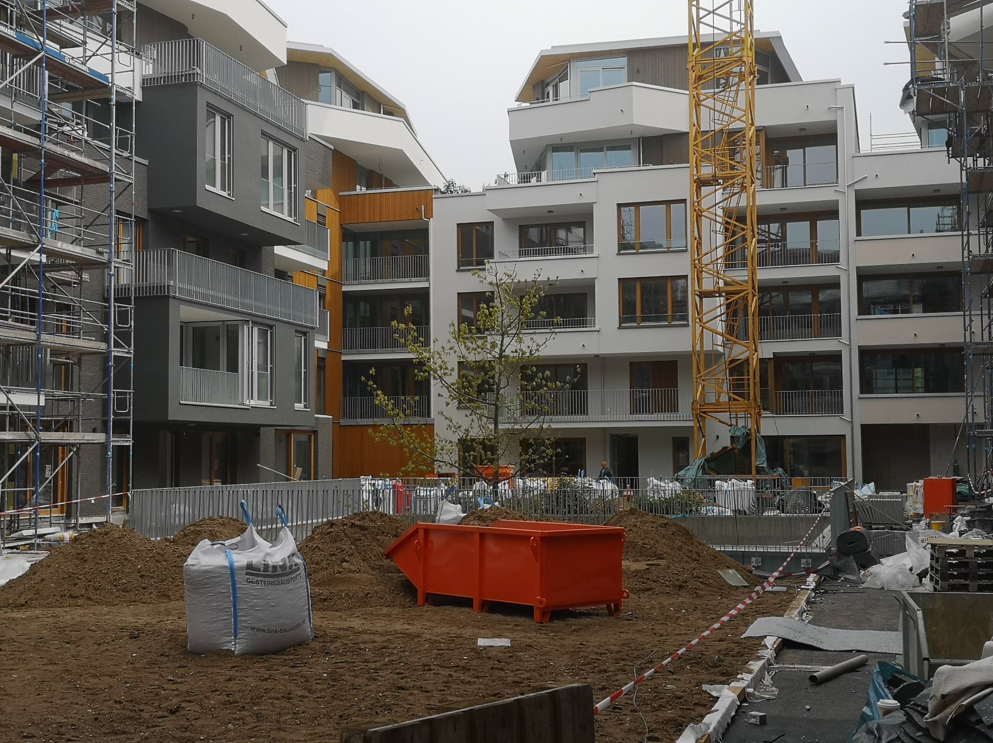 Plac budowy z widokiem na nowoczesne budynki mieszkalne w trakcie budowy, widoczne rusztowania, dźwig, kontener na gruz i worki z materiałami budowlanymi.