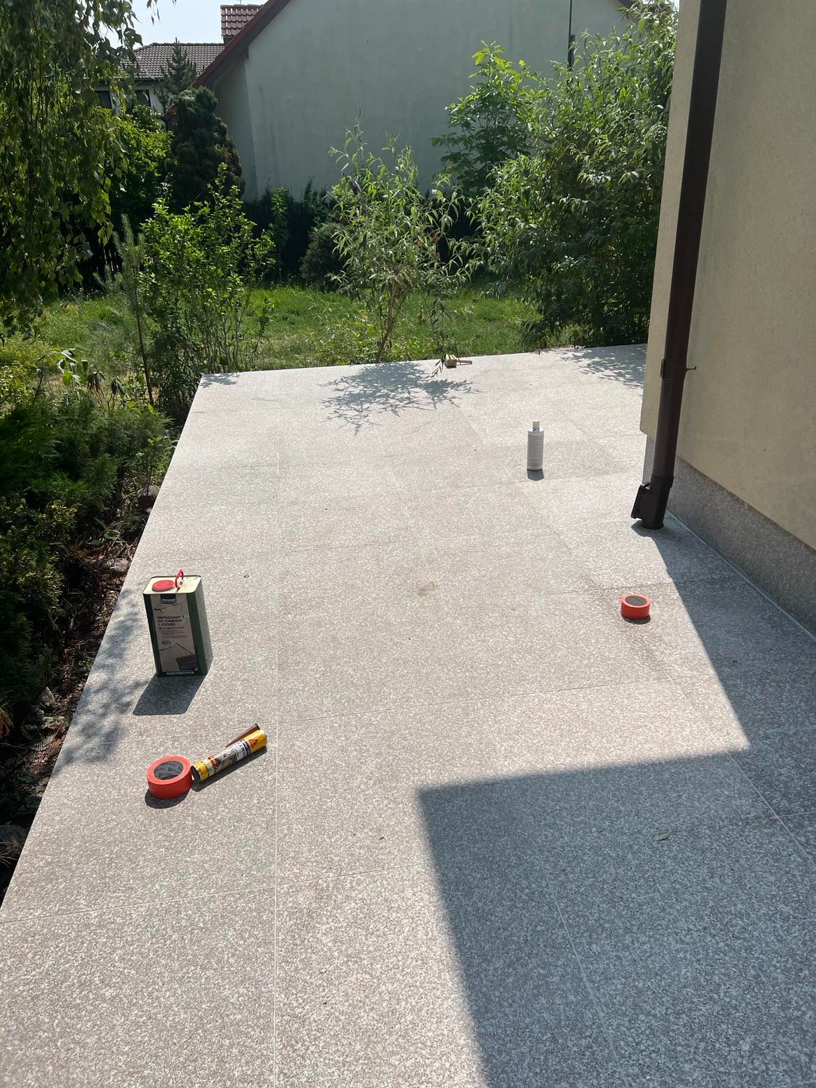 Szary taras z płytek po impregnacji, widoczne narzędzia i preparaty do konserwacji, w tle budynek i zieleń.