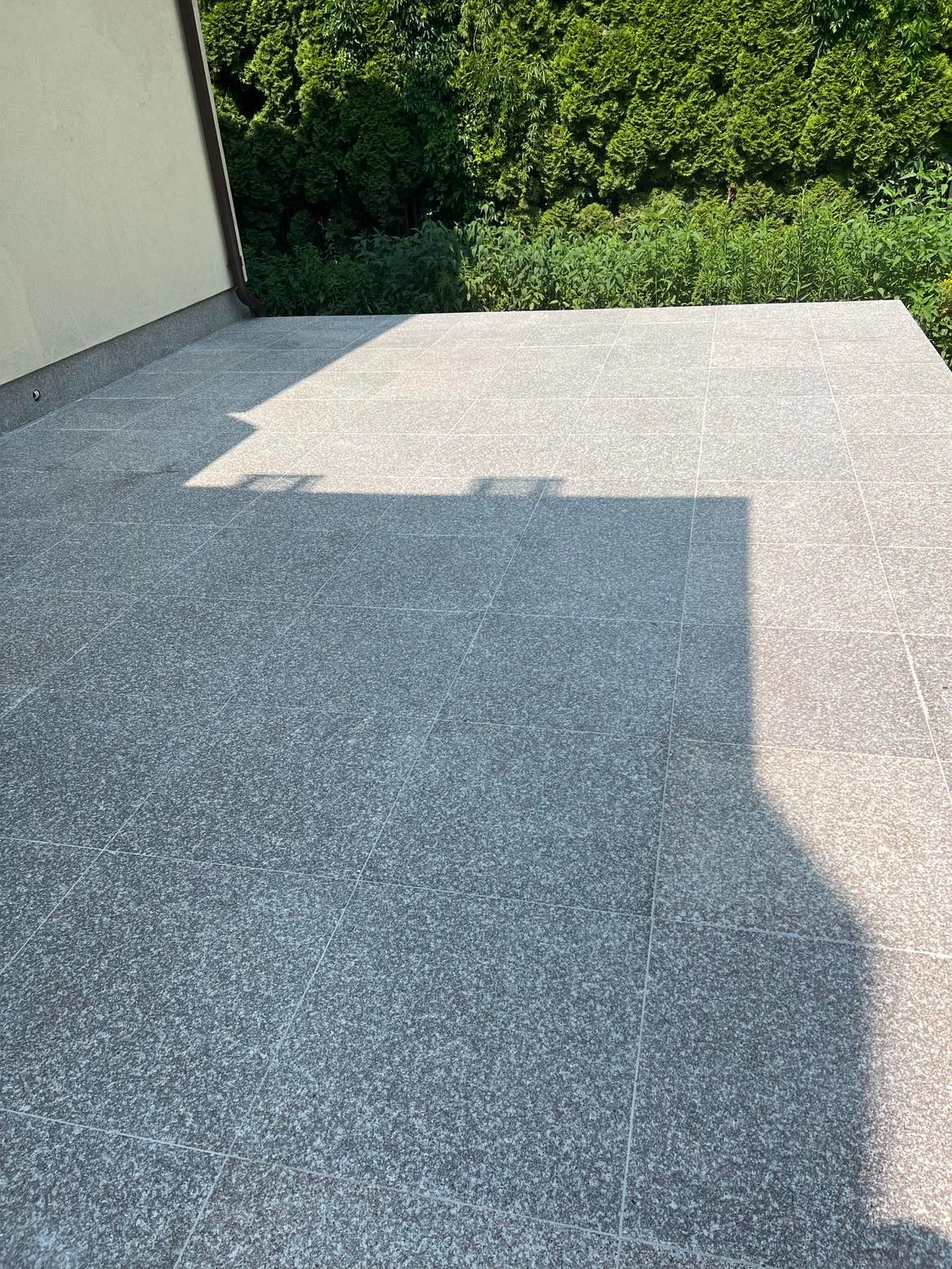 Granitowa posadzka tarasu w odcieniach szarości, z widocznym cieniem rzucanym przez element konstrukcyjny budynku i zielenią w tle.