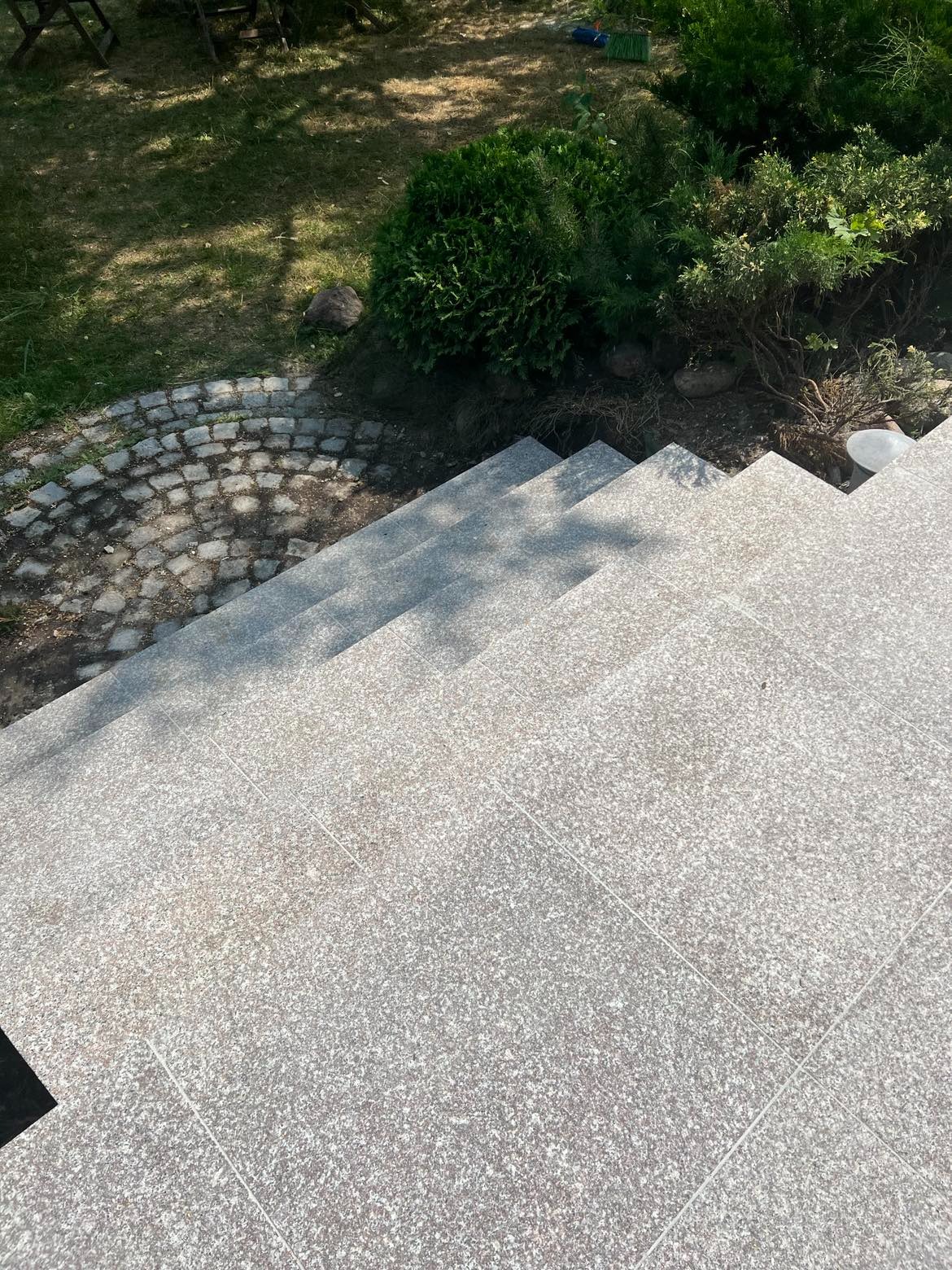 Zewnętrzne schody wyłożone szarymi płytami granitowymi, prowadzące z ogrodu z kostką brukową i zielenią.