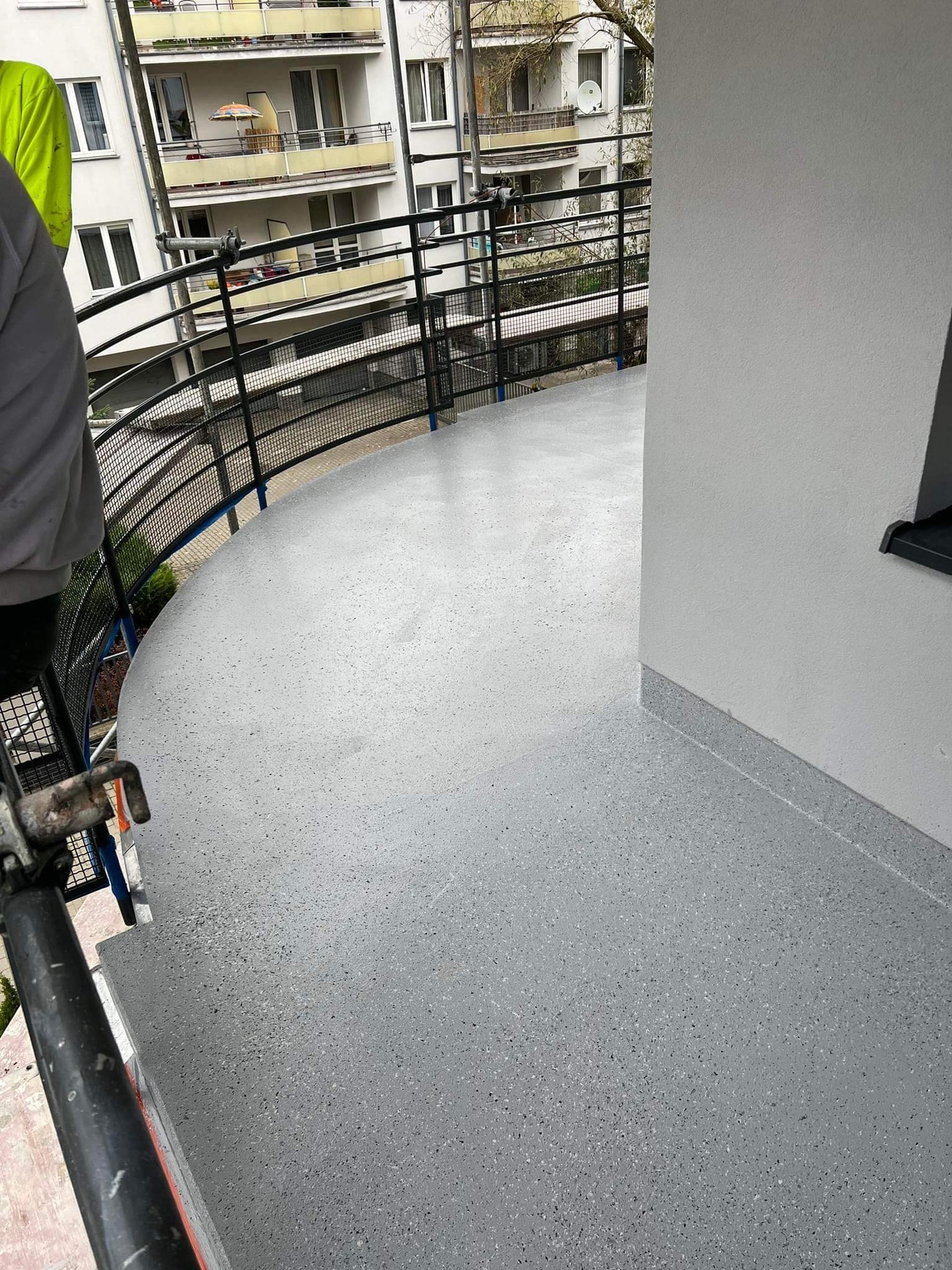 Świeżo wylana, gładka, szara posadzka żywiczna na balkonie z czarną metalową balustradą, widok z rusztowania, budynek mieszkalny w tle.