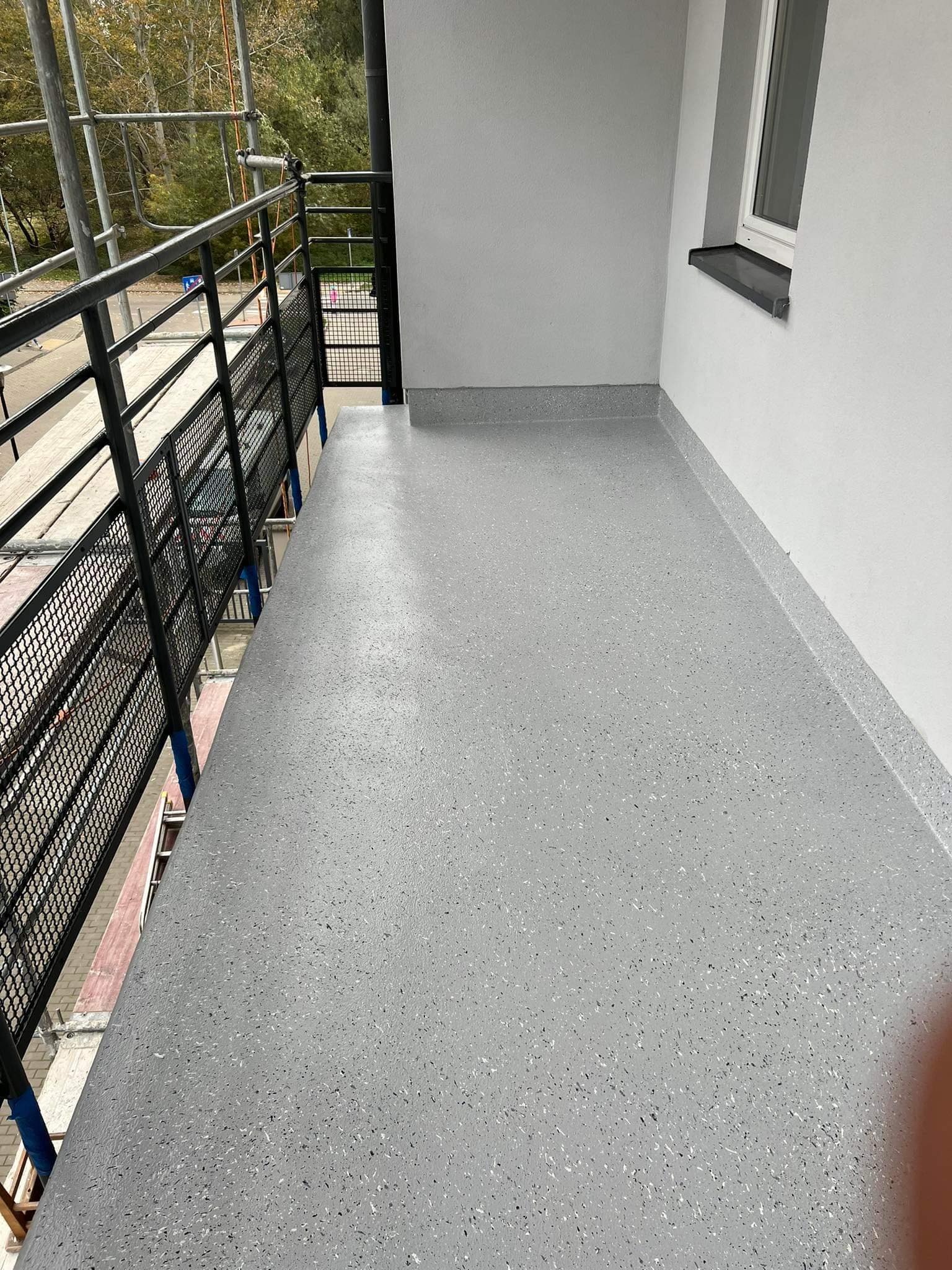 Szary balkon z nową, gładką posadzką żywiczną z delikatnym wzorem, widoczne metalowe barierki i fragment rusztowania.