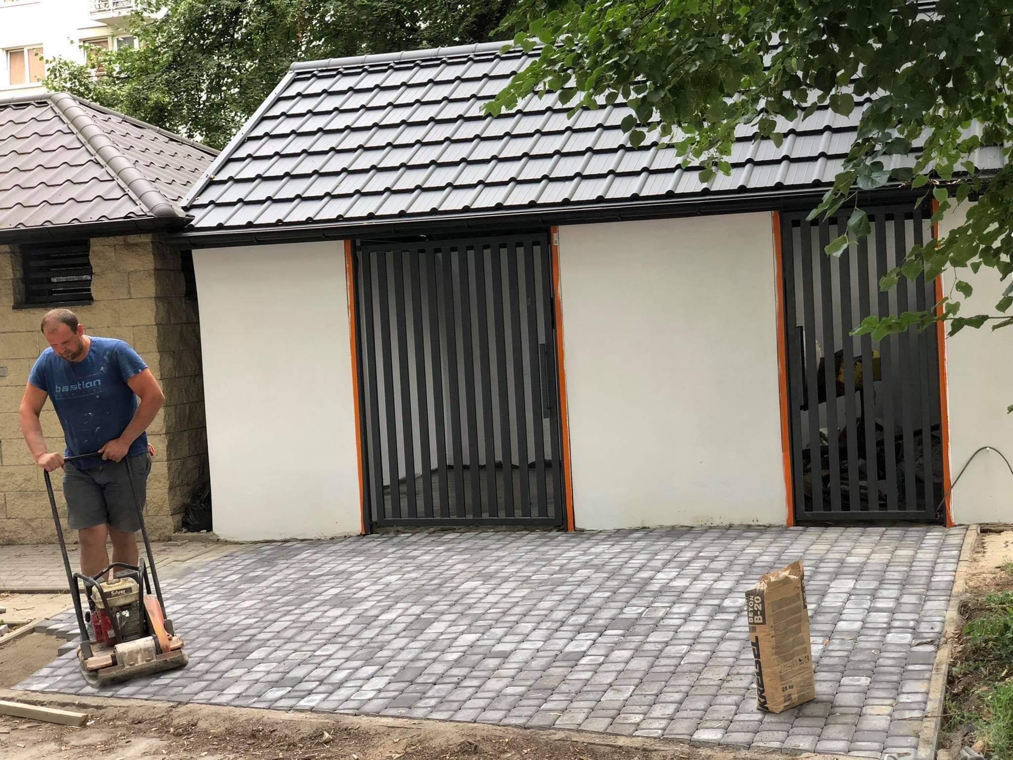 Pracownik zagęszcza nowo ułożoną kostkę brukową na podjeździe, w tle budynek z białymi ścianami i czarnymi, metalowymi bramami oraz widoczne drzewa.
