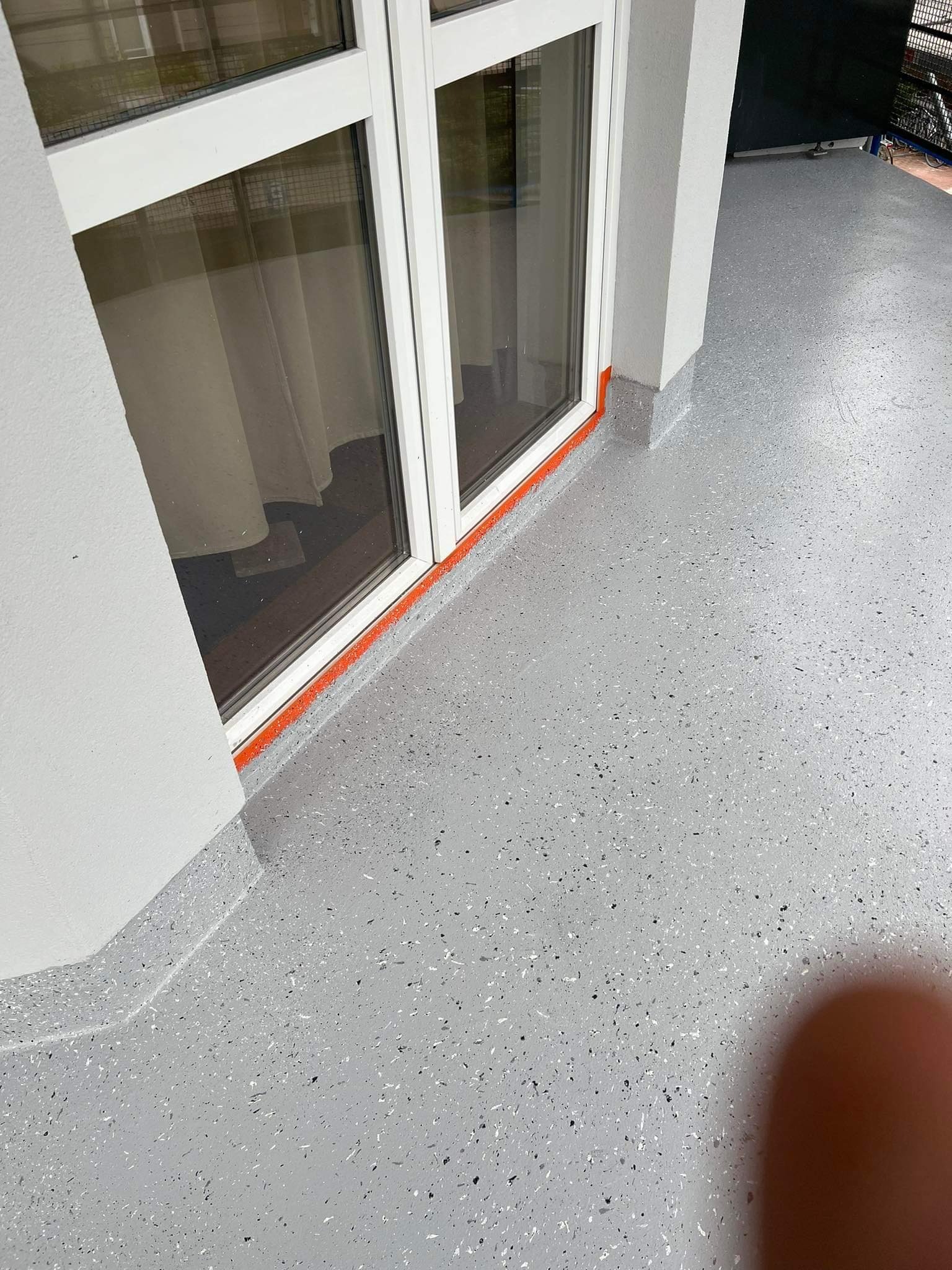 Szara, epoksydowa posadzka balkonu z białymi i czarnymi drobinkami, zabezpieczona pomarańczową taśmą malarską przy białych, przeszklonych drzwiach balkonowych.