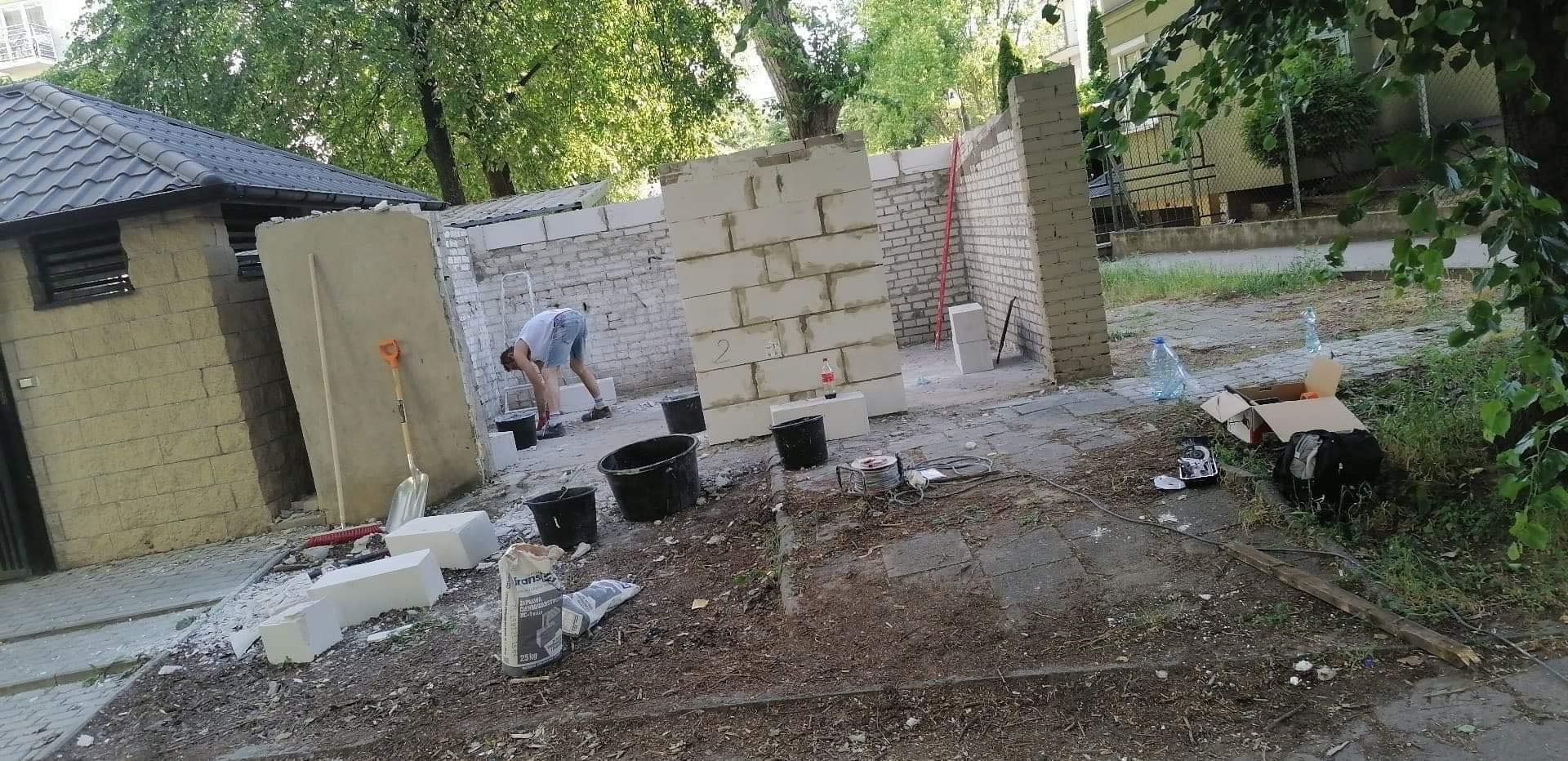 Budowa ścian z bloczków betonowych na zewnątrz budynku, widoczne narzędzia i materiały budowlane, pracownik w tle.