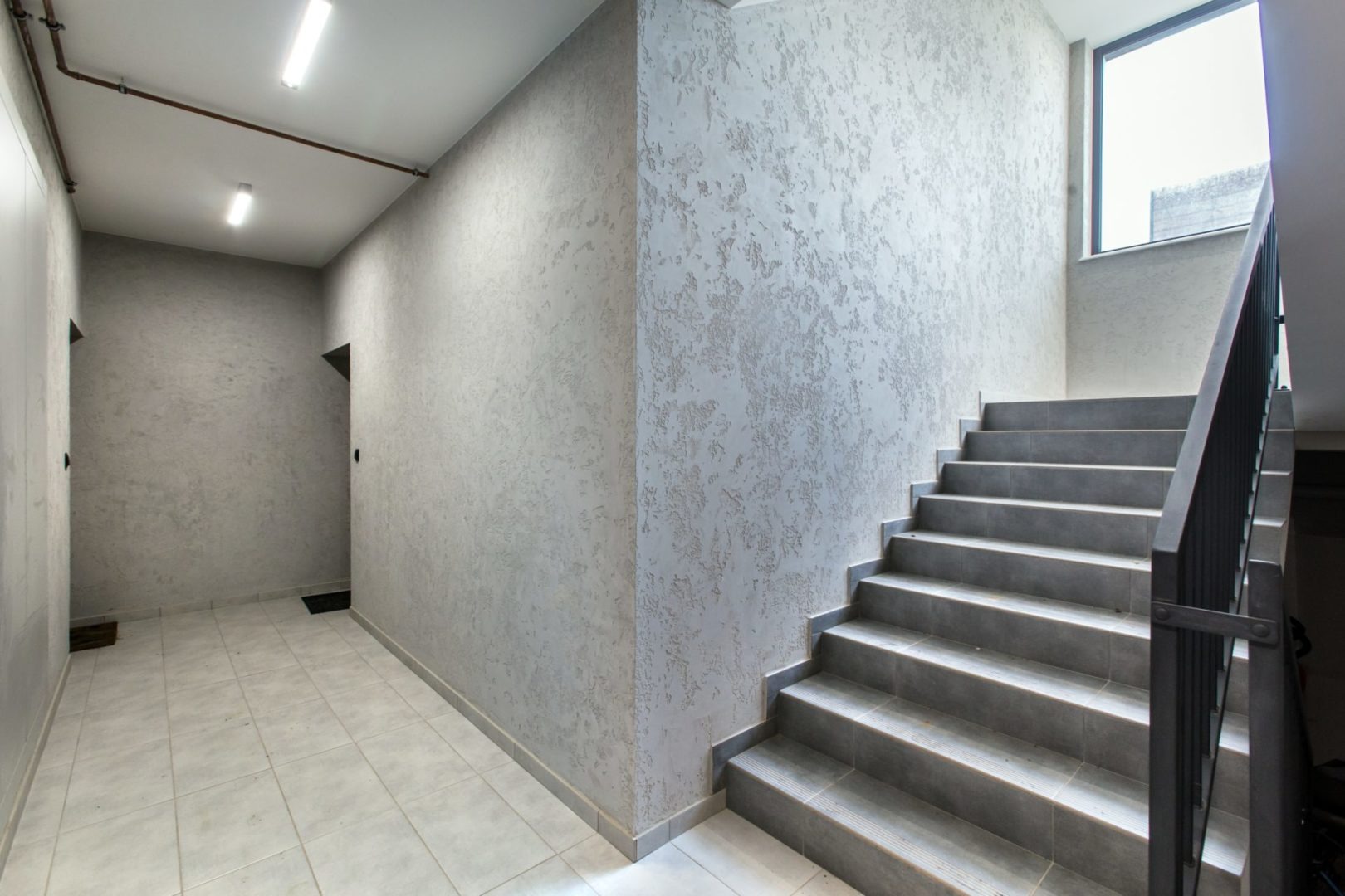 Korytarz w biurowcu po remoncie, ściany pokryte szpachlą dekoracyjną w odcieniach szarości, widoczne schody z szarymi płytkami i czarną balustradą, biała podłoga z płytek, oświetlenie sufitowe.