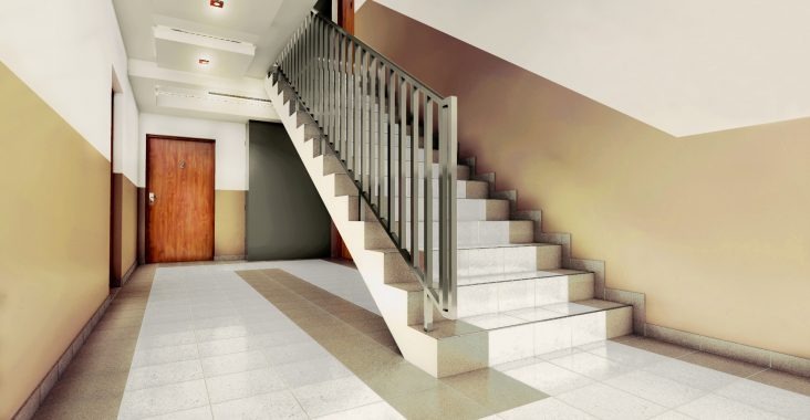 Korytarz w bloku z pomalowanymi ścianami w dwóch kolorach, białe płytki na podłodze i schody z metalową barierką.