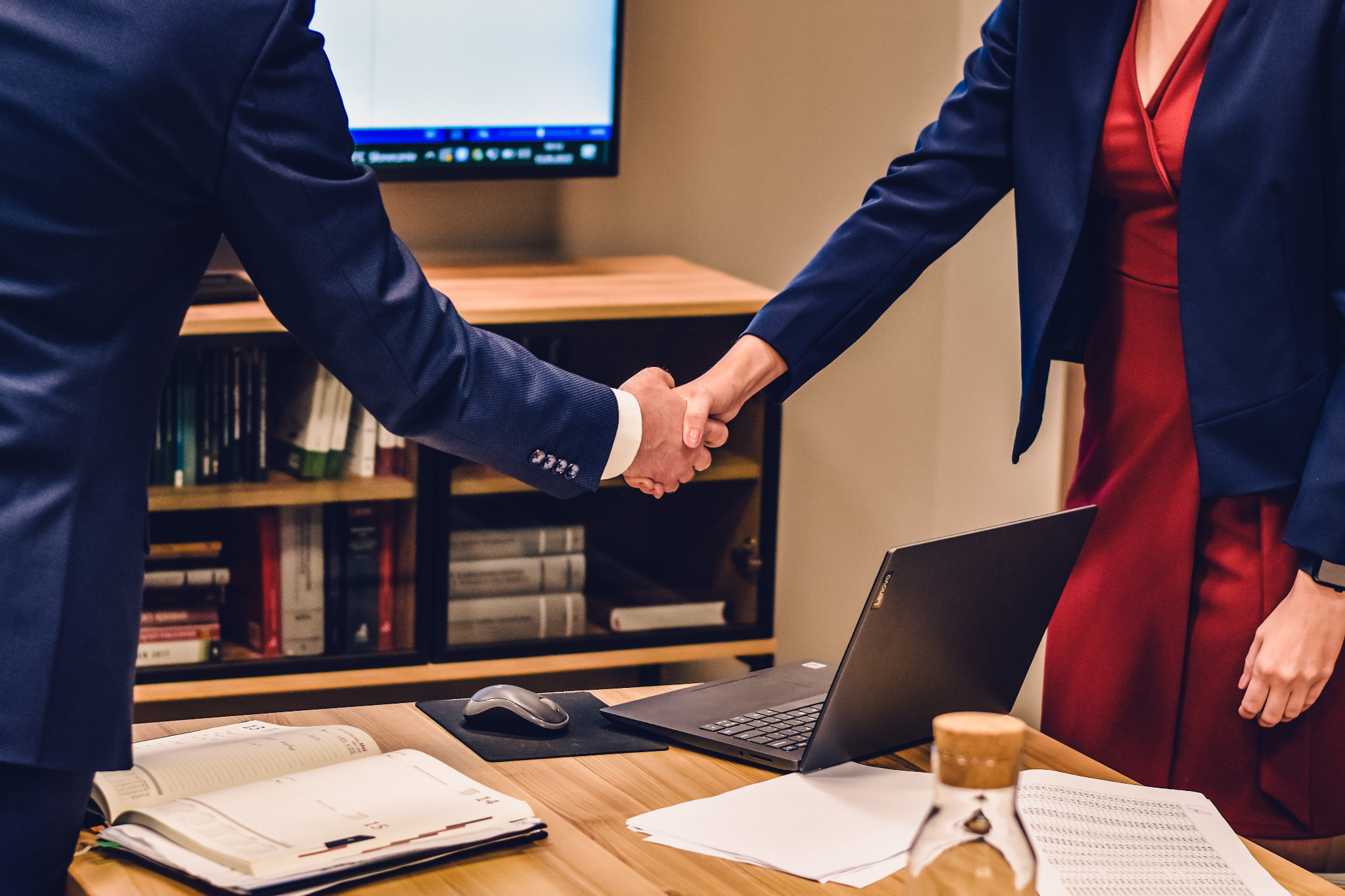 Uścisk dłoni nad biurkiem z laptopem, kalendarzem i dokumentami, w tle regał z książkami i monitor.