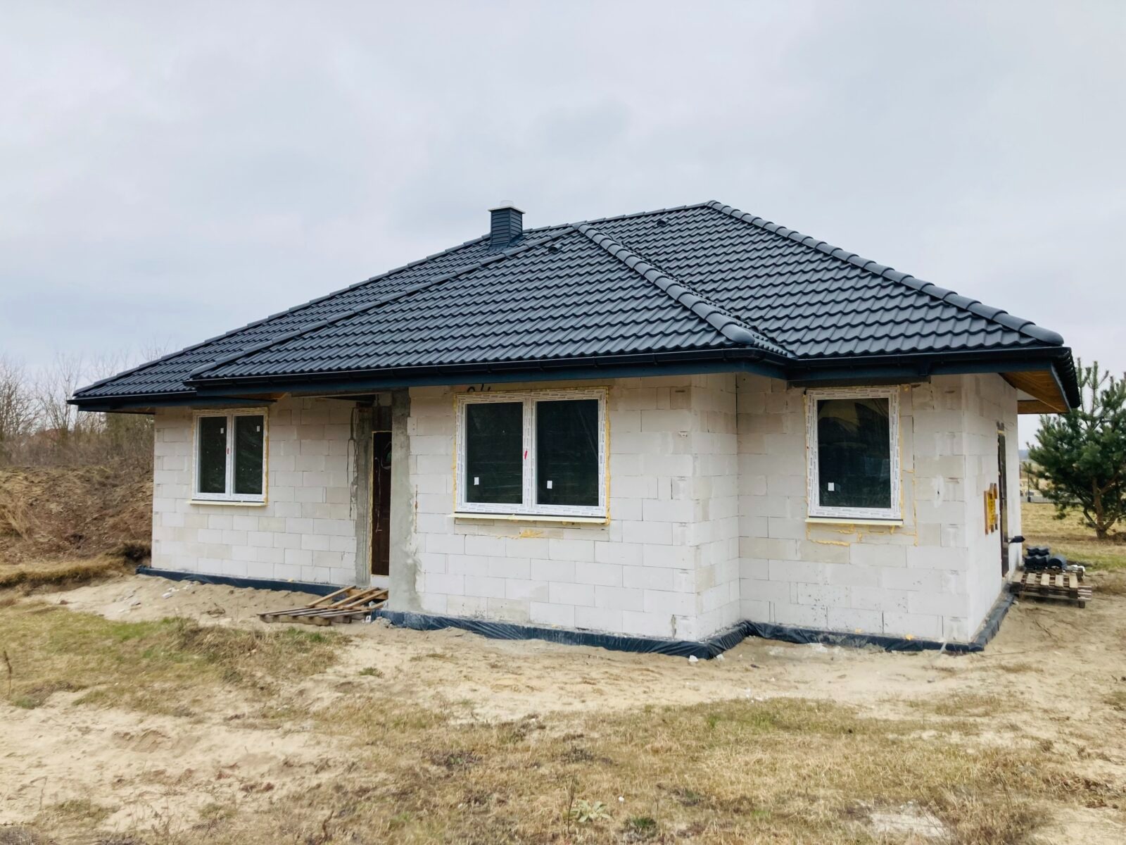 Niedokończony dom murowany z białych bloczków, czarny dach z dachówki ceramicznej, białe okna z folią ochronną, widoczne fundamenty zabezpieczone folią, teren wokół domu piaszczysty.