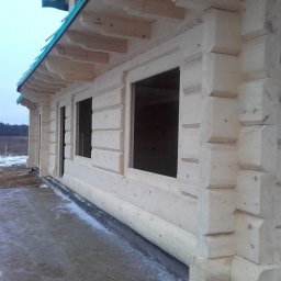 Domki z Mazur - Surowy stan domu z bali: widok z boku na ściany z otworami okiennymi, zadaszenie z zieloną folią i resztki śniegu na ziemi. Widoczne detale łączeń bali.