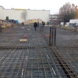 Domki z Mazur - Zbrojenie fundamentu domu jednorodzinnego z widocznymi prętami pionowymi, przygotowane do zalania betonem. W tle budynki i drzewa.