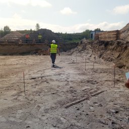 Domki z Mazur - Przygotowanie terenu pod budowę: wykop z wyznaczonym obrysem fundamentów, pracownicy w odblaskowych kamizelkach i paliki geodezyjne. W tle składowane drewno.