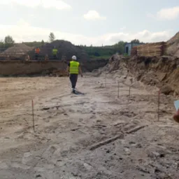 Przygotowanie terenu pod budowę: wykop z wyznaczonym obrysem fundamentów, pracownicy w odblaskowych kamizelkach i paliki geodezyjne. W tle składowane drewno.