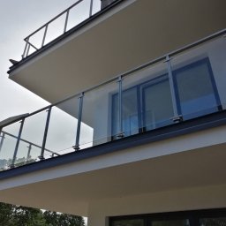Domki z Mazur - Nowoczesny balkon z szklaną balustradą i metalowymi elementami, widok z dołu, zieleń drzew w tle. Minimalistyczny design, jasna kolorystyka.