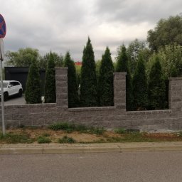 Domki z Mazur - Nowoczesny mur z szarej cegły z wkomponowanymi tujami, tworzący ogrodzenie posesji z białym autem w tle. Widoczny znak drogowy. Umiarkowane oświetlenie.