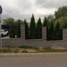 Nowoczesny mur z szarej cegły z wkomponowanymi tujami, tworzący ogrodzenie posesji z białym autem w tle. Widoczny znak drogowy. Umiarkowane oświetlenie.