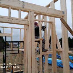 Domki z Mazur - Montera w trakcie budowy domu szkieletowego. Widoczna drewniana konstrukcja, elementy złączone metalowymi łącznikami. Praca na wysokości.