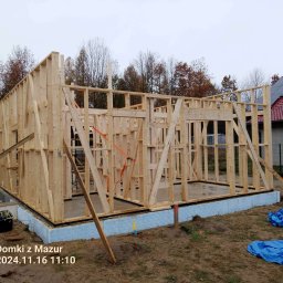 Domki z Mazur - Szkieletowa konstrukcja domu w trakcie budowy, widoczne drewniane elementy ścian i fundamenty z izolacją, na zewnątrz rozłożona folia budowlana.